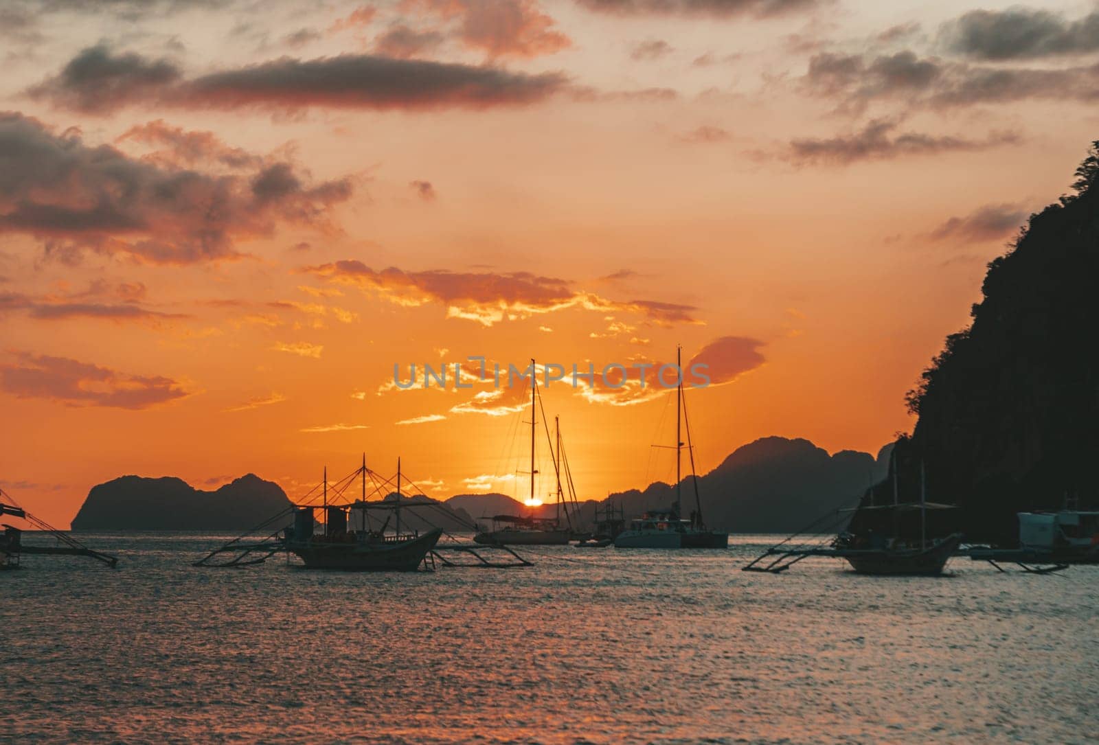 Multiple boats are anchored in a calm bay under a vibrant sunset. The sky glows with shades of orange and gold, while dark clouds create dramatic contrasts.