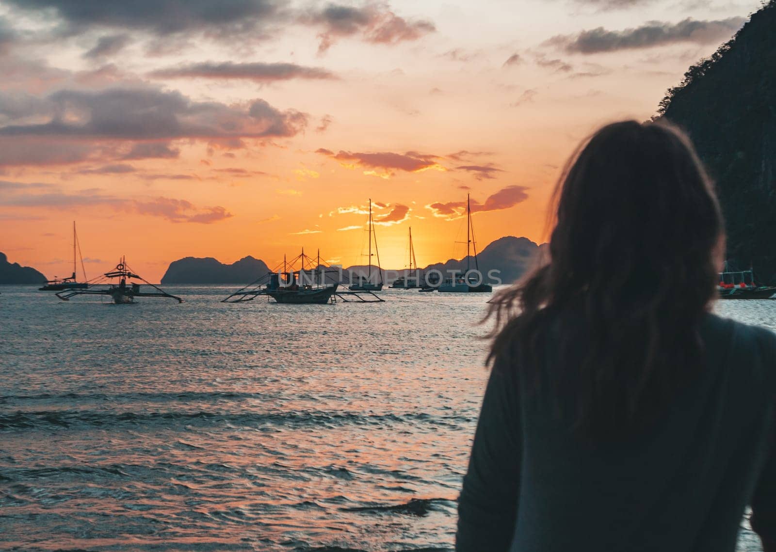 A woman stands on the beach in El Nido, Palawan, watching a stunning sunset over the calm ocean waters, with boats in the distance.