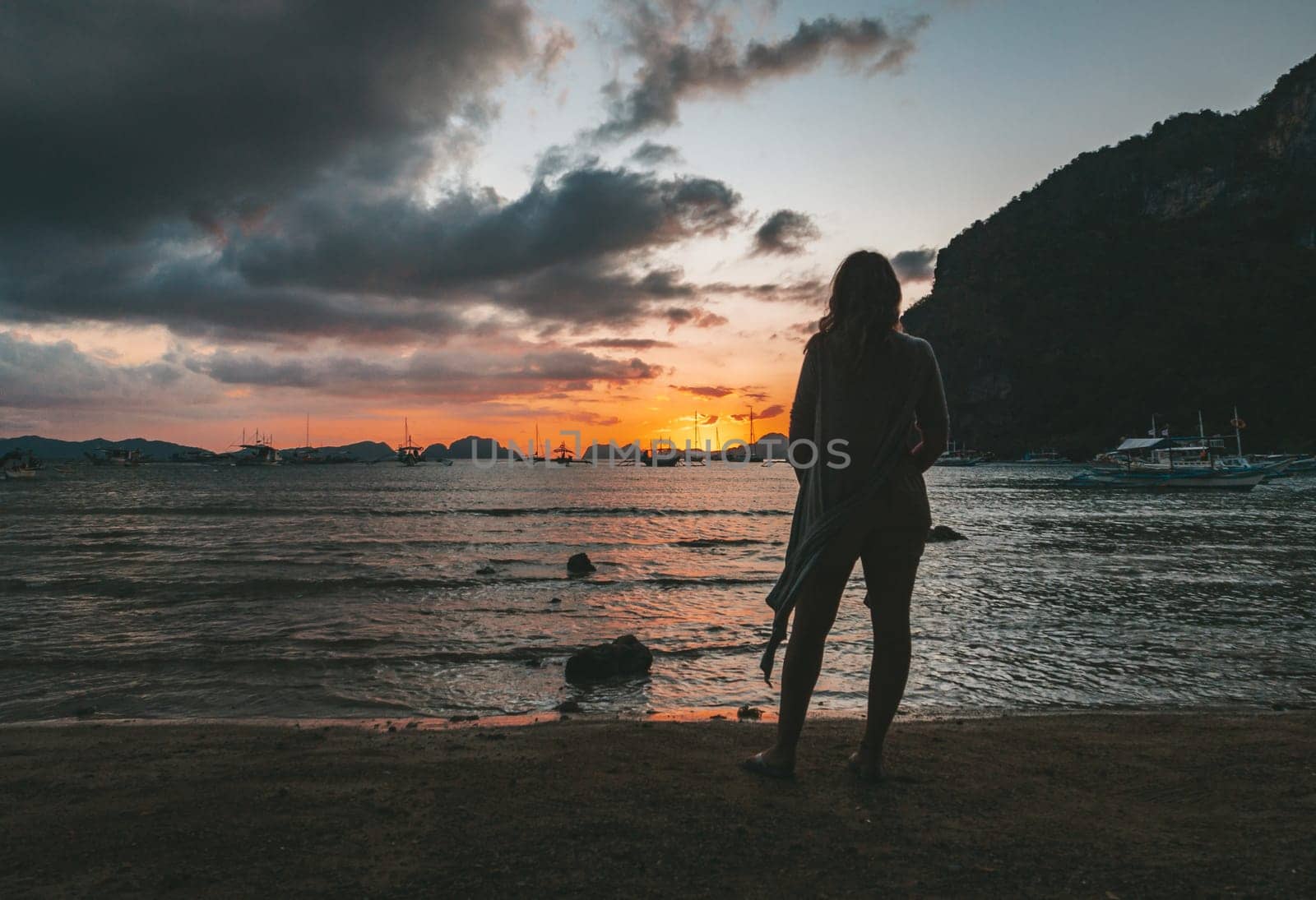 A woman stands on the beach in El Nido, Palawan, watching a stunning sunset over the calm ocean waters, with boats in the distance.