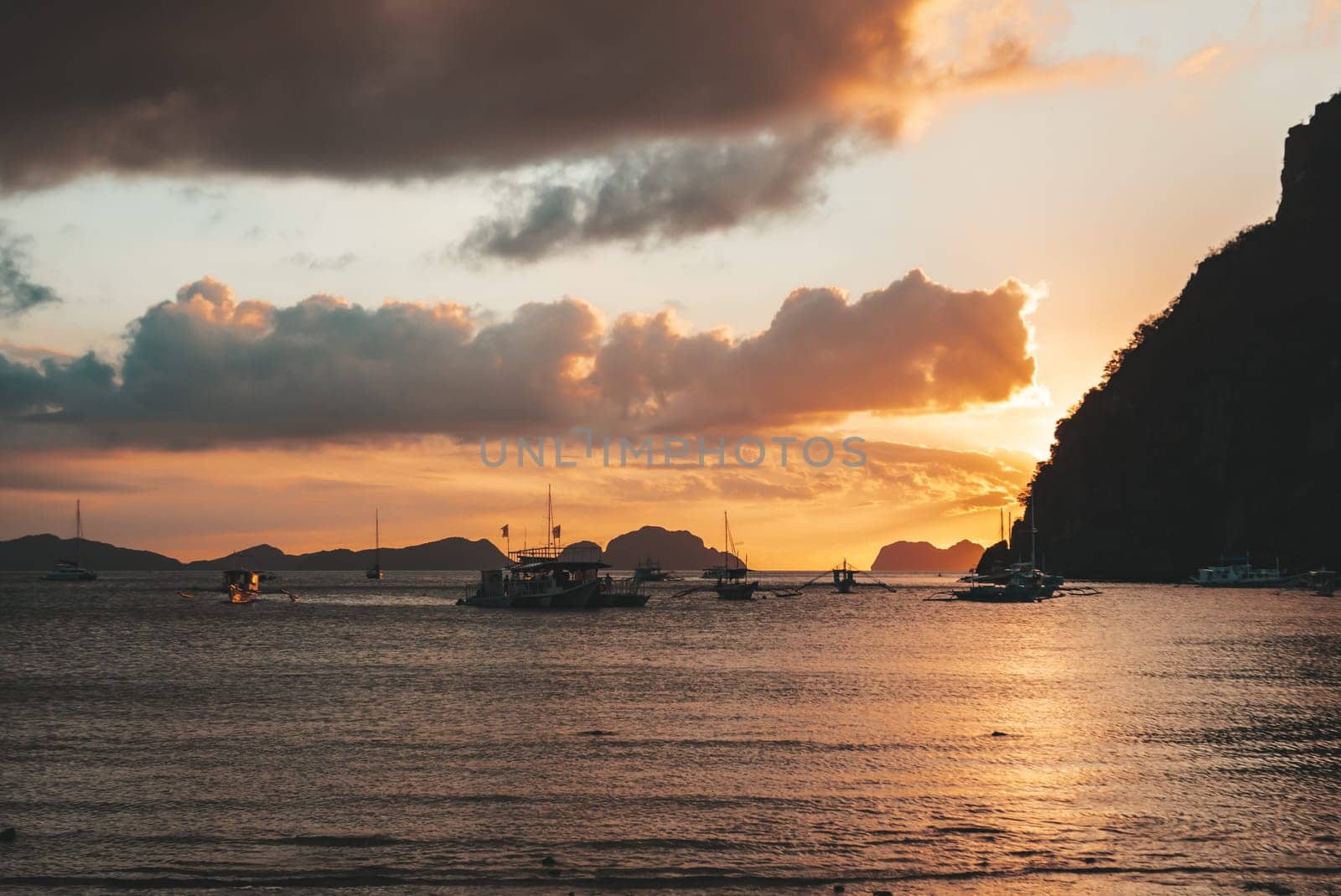 Multiple boats are anchored in a calm bay under a vibrant sunset. The sky glows with shades of orange and gold, while dark clouds create dramatic contrasts.