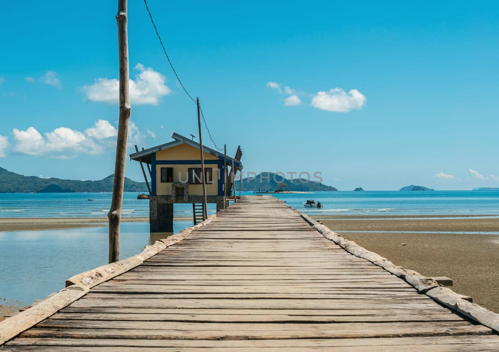 Rustic wooden pier extending towards a small hut situated on the coastline, under a clear blue sky. Scenic mountains are visible in the background along with the calm sea and scattered clouds.