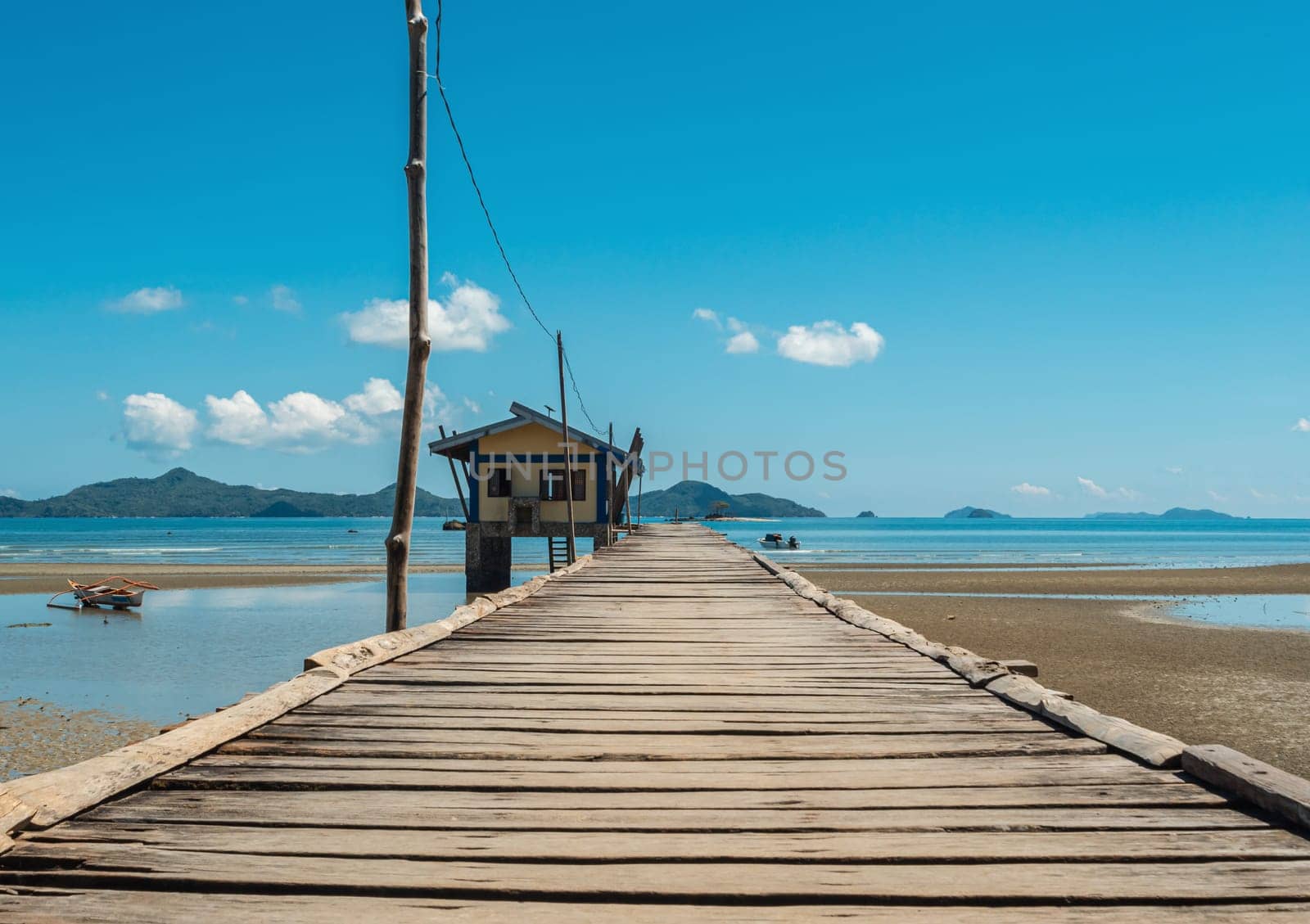 Rustic wooden pier extending towards a small hut situated on the coastline, under a clear blue sky. Scenic mountains are visible in the background along with the calm sea and scattered clouds.