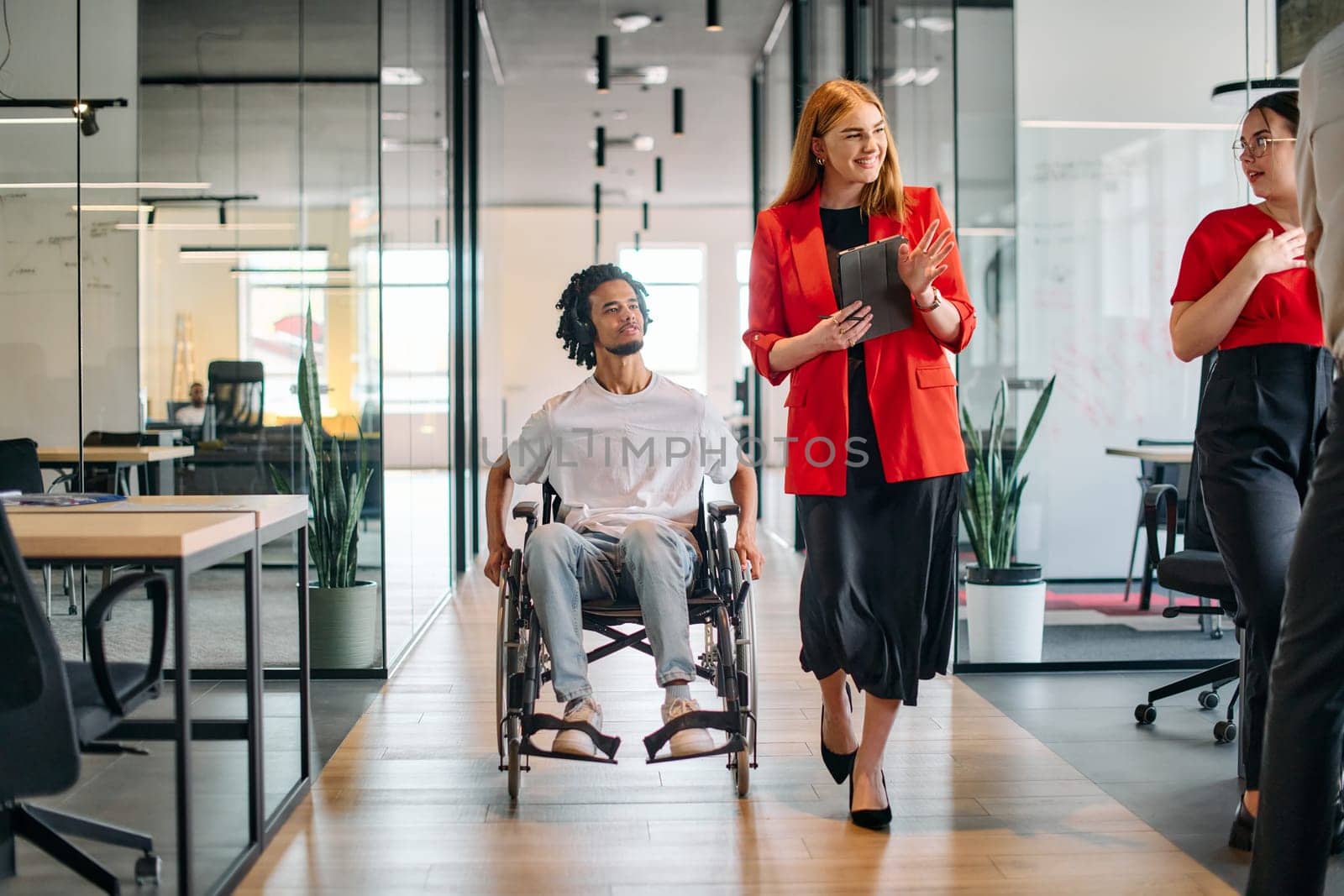 A business leader with her colleague, an African-American businessman who is a disabled person, pass by their colleagues who work in modern offices.