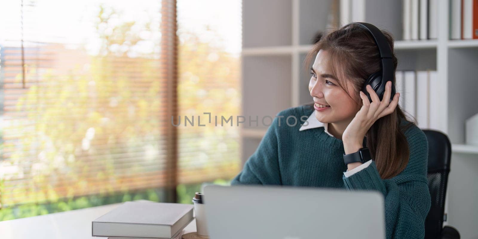 Young woman smiling while wearing headphones and using a laptop in a bright home office.