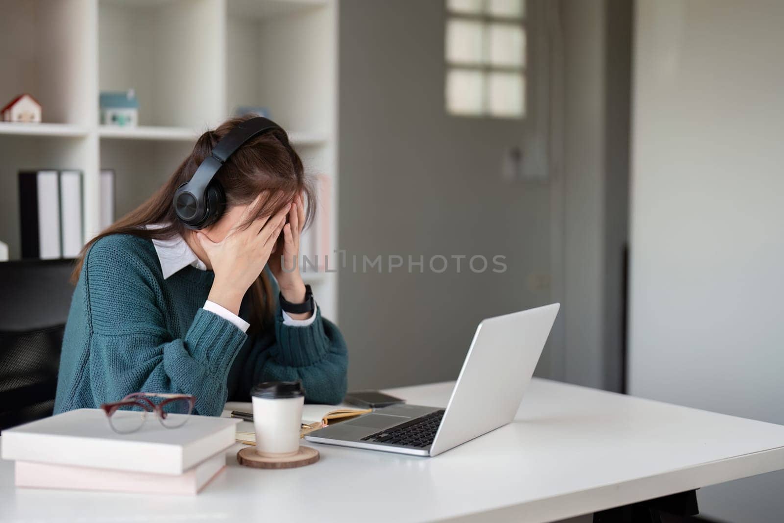 Young woman feeling stressed while wearing headphones and using a laptop in a home office.