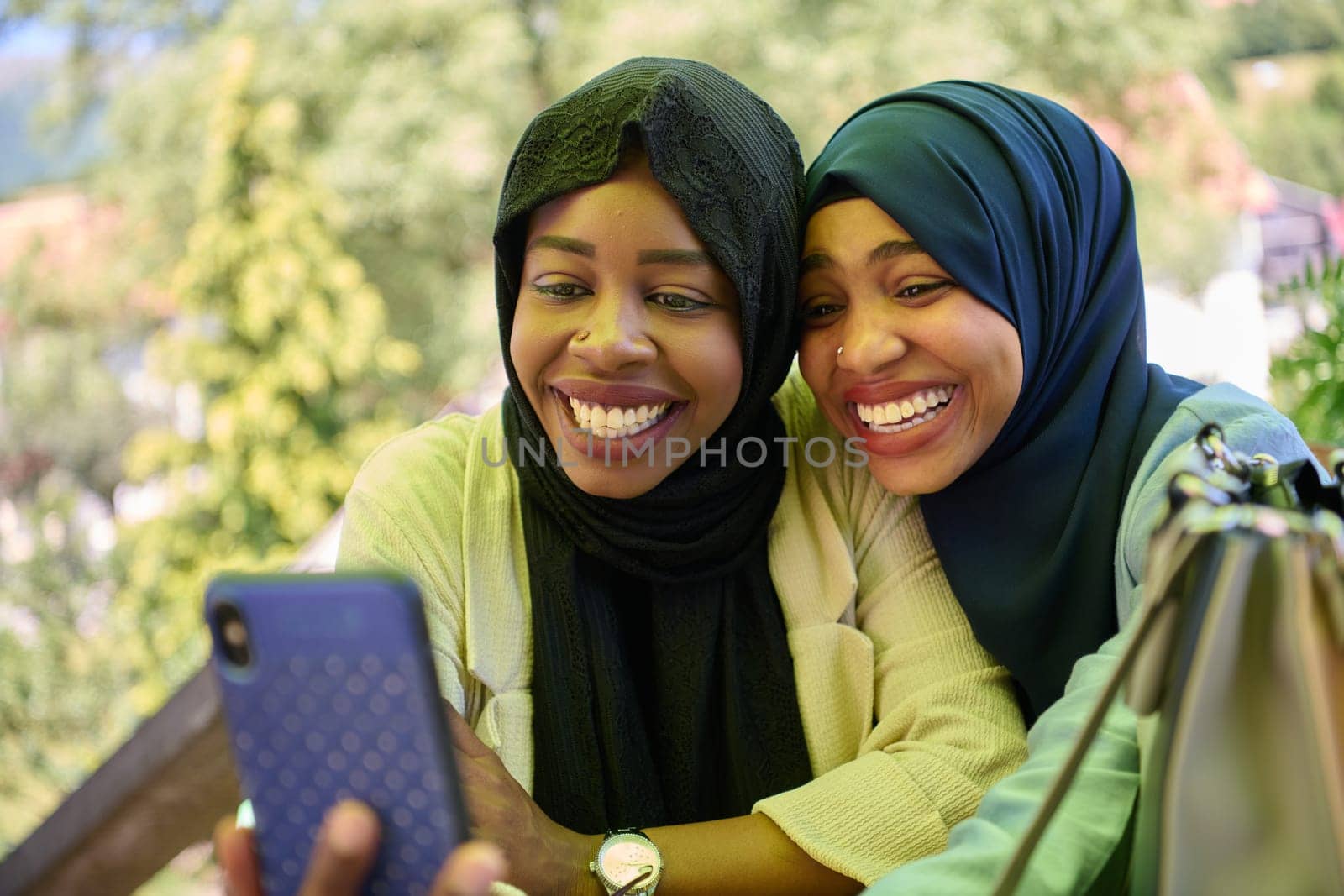 Two Middle Eastern Muslim women, adorned in hijabs, capture a moment of friendship and joy as they take selfies on a smartphone while seated in a natural setting.