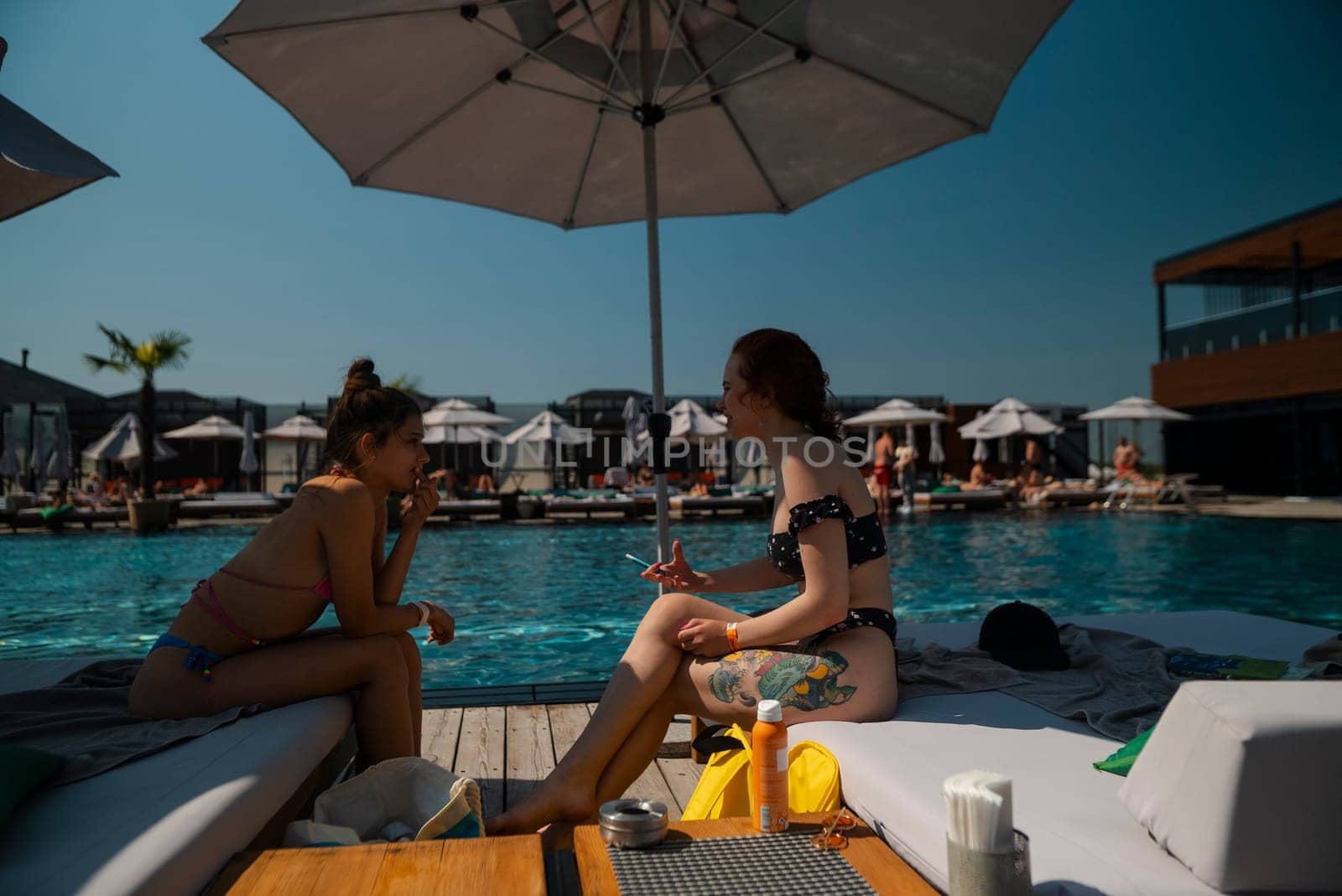 A pair of youthful girlfriends relax by the pool, soaking up the sun in their bikinis. High quality photo