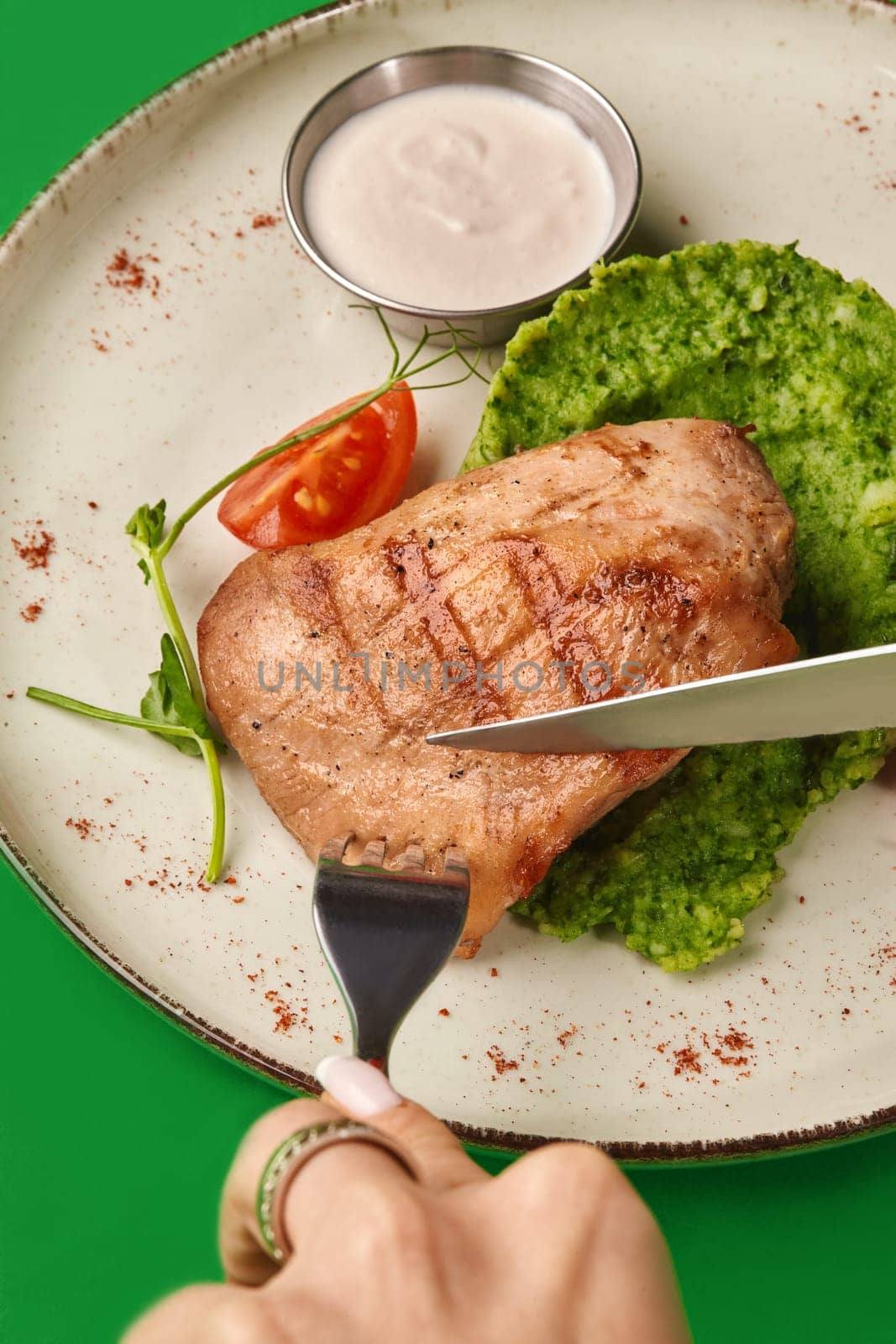 Close-up of female hands slicing grilled chicken breast served on bed of broccoli puree with side of creamy sauce and cherry tomato on vibrant green background