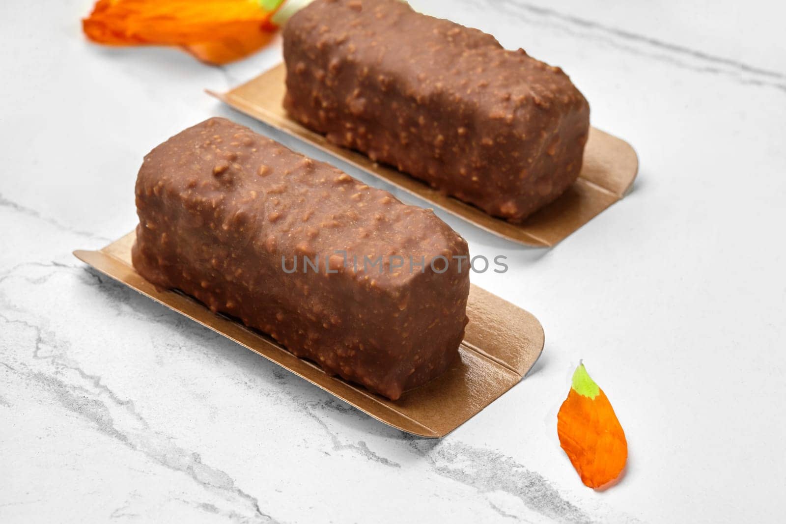 Milk chocolate-glazed cottage cheese bars sprinkled with nuts, presented on golden cardboards with marble background and orange flower petals