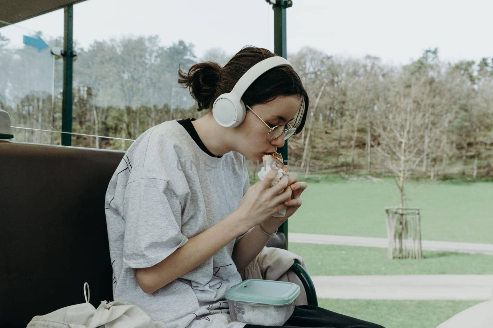 Portrait of one beautiful Caucasian teenage girl in white headphones eating, biting a sandwich, sitting sideways in the open carriage of a tourist train traveling through the reserve on a spring day, side view close-up.