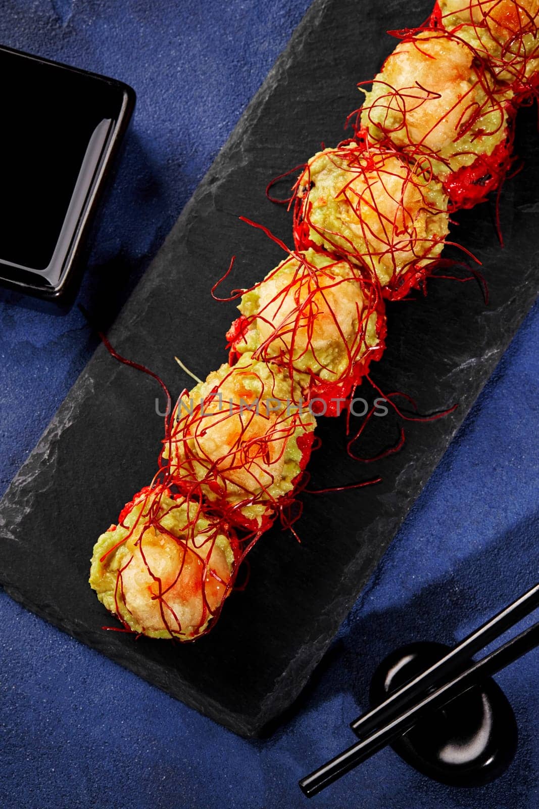 Red tobiko sushi rolls topped with avocado spread and shrimp tempura garnished with spicy togarashi threads traditionally served with soy sauce on black slate board on blue backdrop. Japanese cuisine