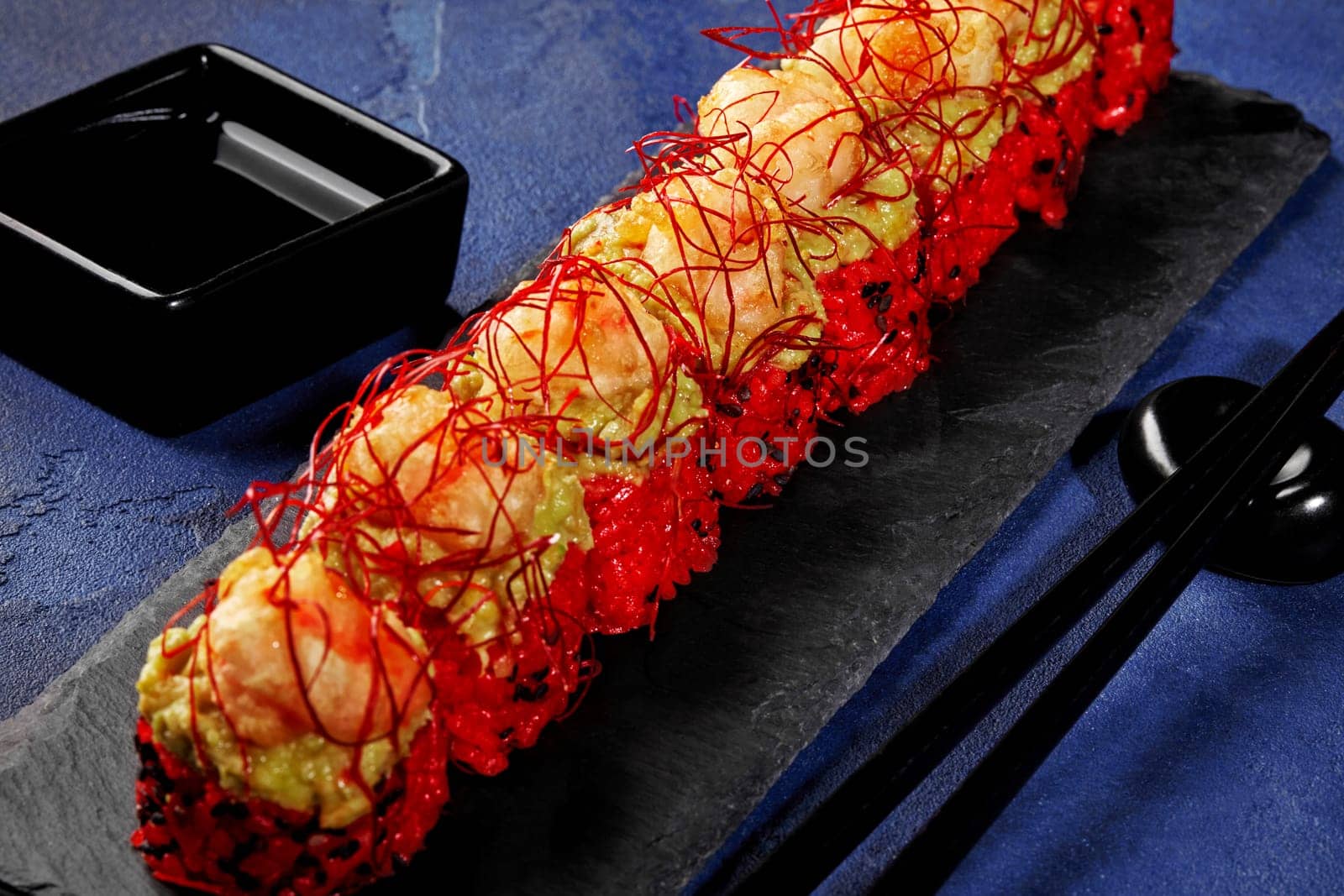 Red rice sushi rolls topped with avocado spread and shrimp tempura sprinkled with spicy chili pepper threads, presented on black slate board with soy sauce on blue background. Japanese cuisine