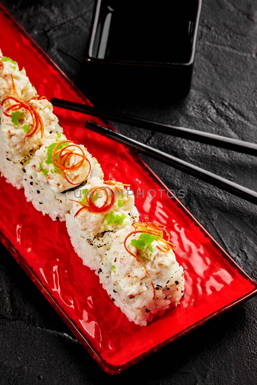 Spicy set of sushi rolls with creamy crab toppings garnished with green wasabi tobiko and fresh chili pepper shavings on red dish, accompanied by soy sauce and chopsticks on textured black background