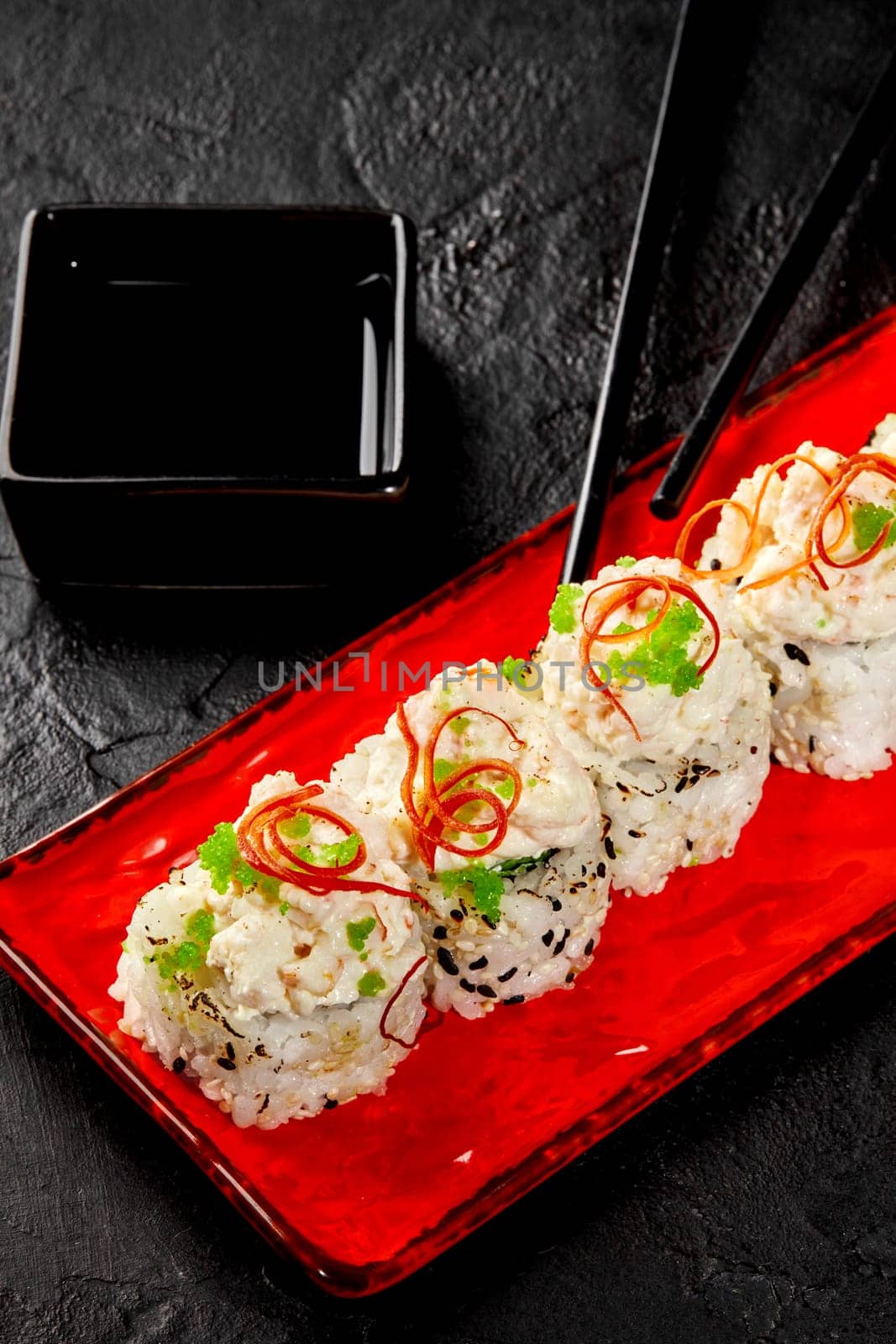 Exquisite sesame coated sushi rolls, topped with creamy seafood caps, green tobiko and chili pepper shavings served on red plate with soy sauce, on black textured surface. Japanese culinary cultures