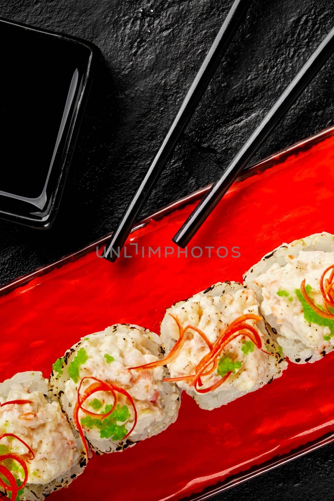 Top view of vibrant red plate with lightly seared sushi rolls with creamy crab toppings, green tobiko and spicy chili shavings, presented against textured black surface with bowl of soy sauce