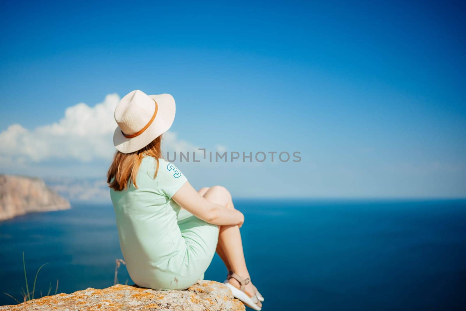 A woman wearing a straw hat is sitting on a rock overlooking the ocean. The sky is clear and blue, and the water is calm. Concept of relaxation and tranquility