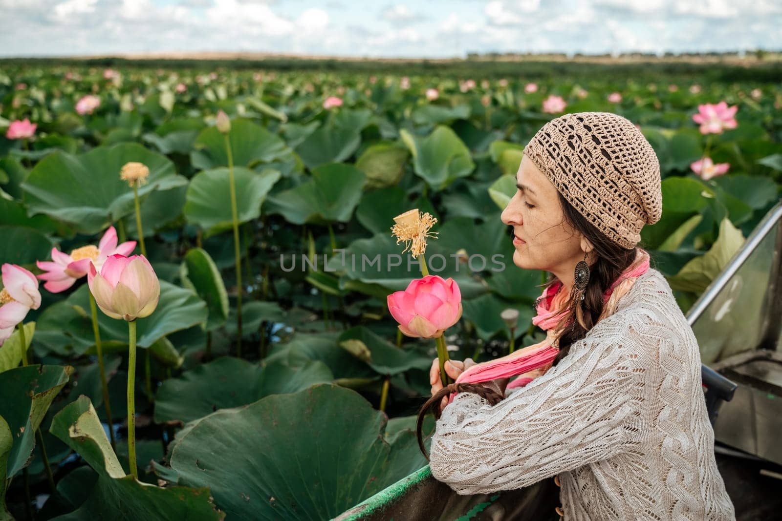 A woman is sitting in a boat in a field of pink lotus flowers. She is wearing a hat and a scarf. by Matiunina