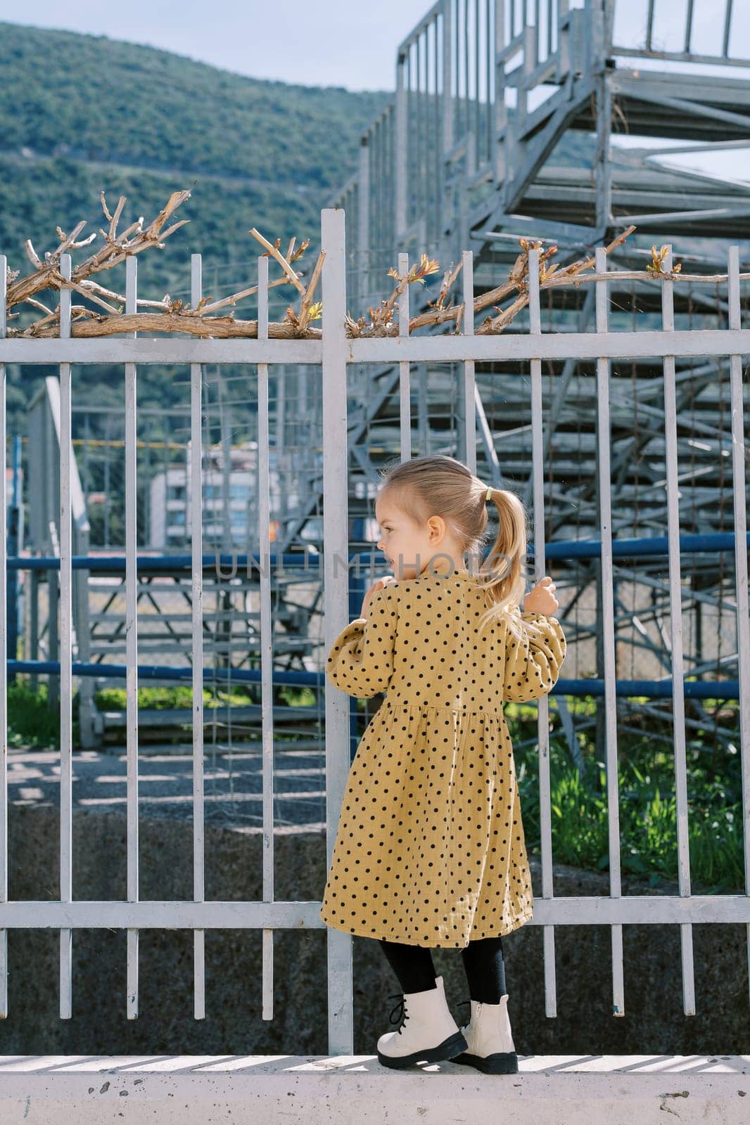 Little girl stands on the base of a metal fence, holding onto the bars and looking to the side. Back view. High quality photo