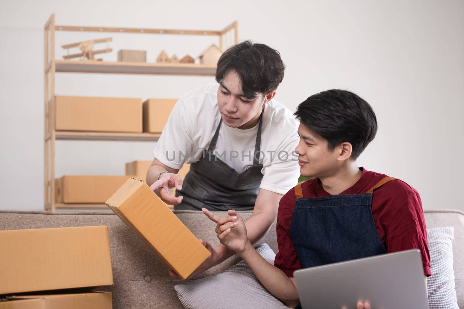 A gay couple running a small business takes order on a laptop and sells product online together in a room full of boxes. by wichayada