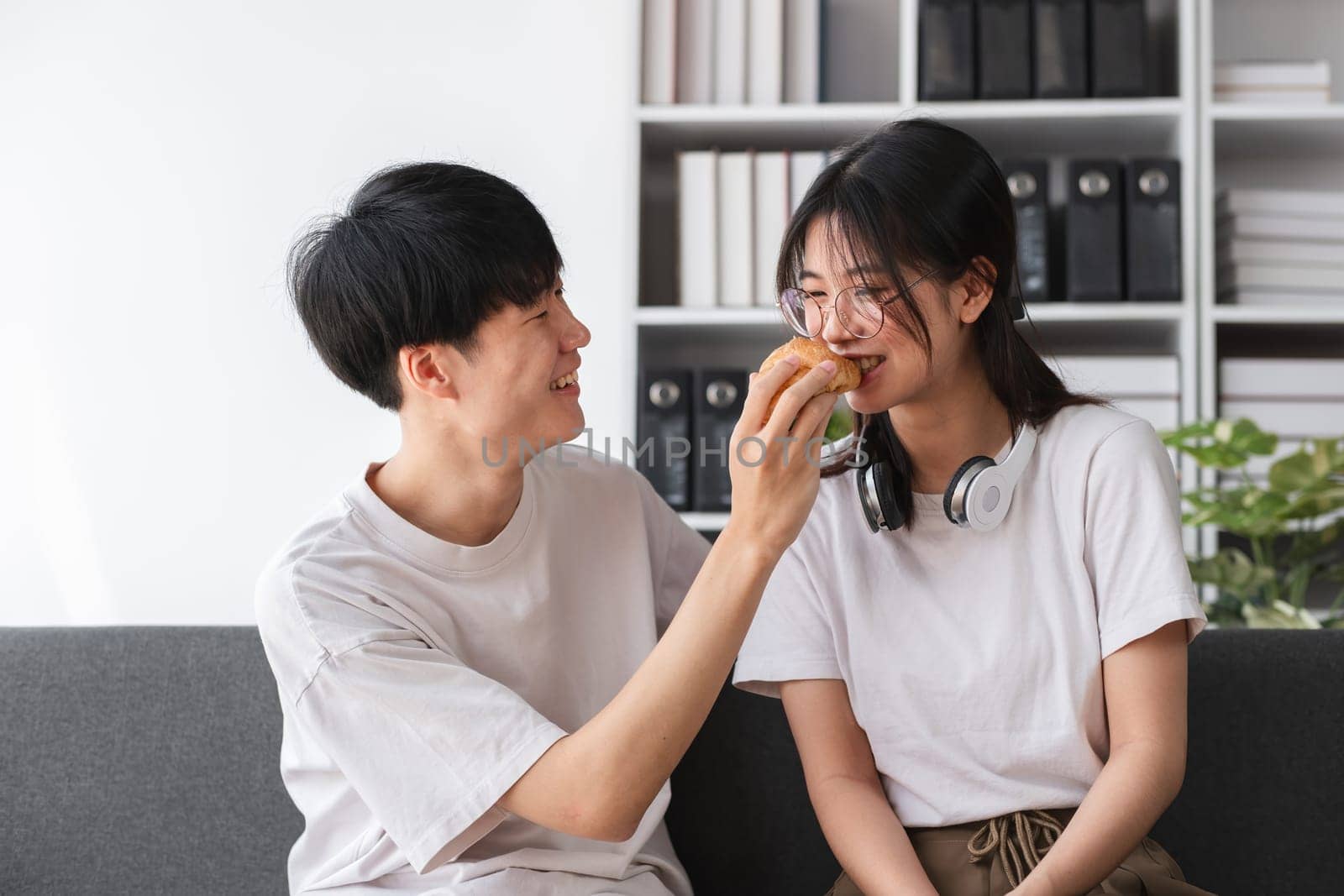 Young couple sitting together on the sofa in the living room Have coffee and snacks together. by wichayada