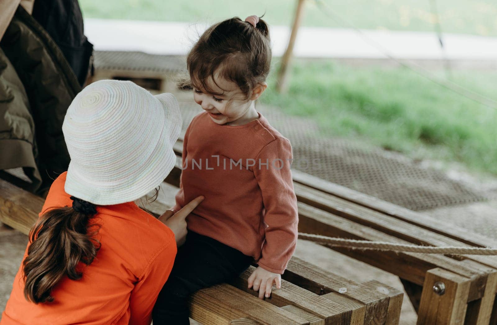 Portrait of two little beautiful Caucasian girls sisters having fun and cute playing with each other and sitting on a wooden bench on a spring day in a park in Belgium, close-up side view from above.