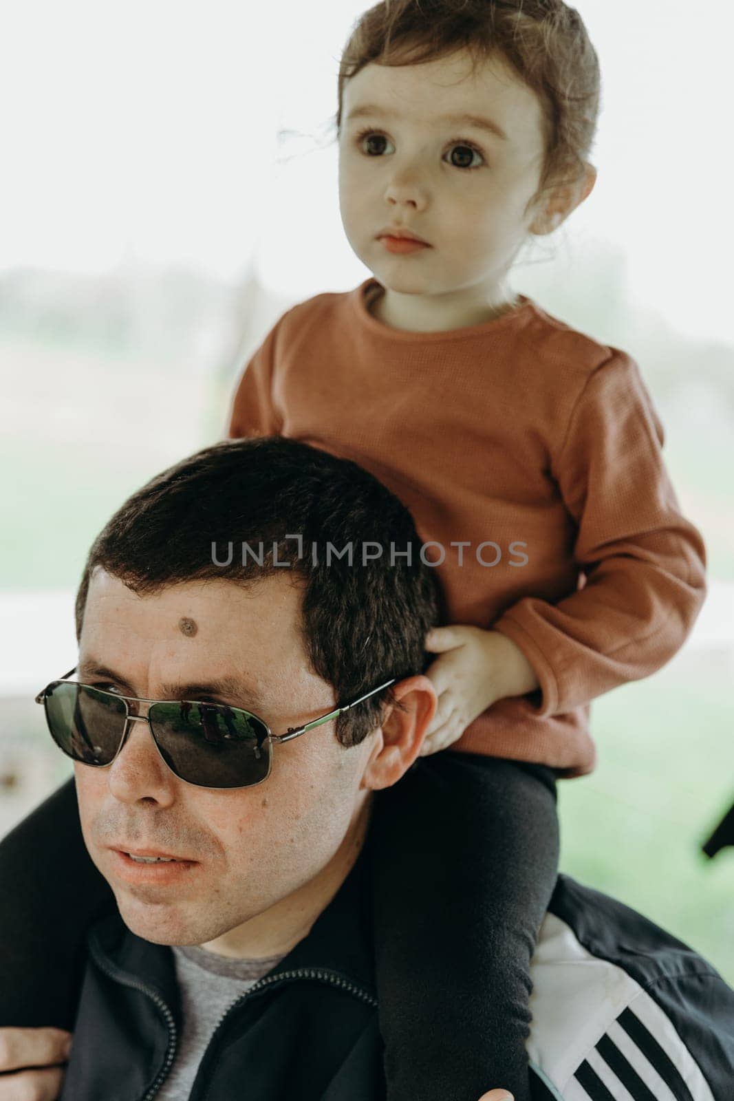 Portrait of one handsome young Caucasian male father in sunglasses holding his little daughter on his shoulders and both looking carefully to the side with interest while walking on a sunny spring day in a public park, close-up side view.