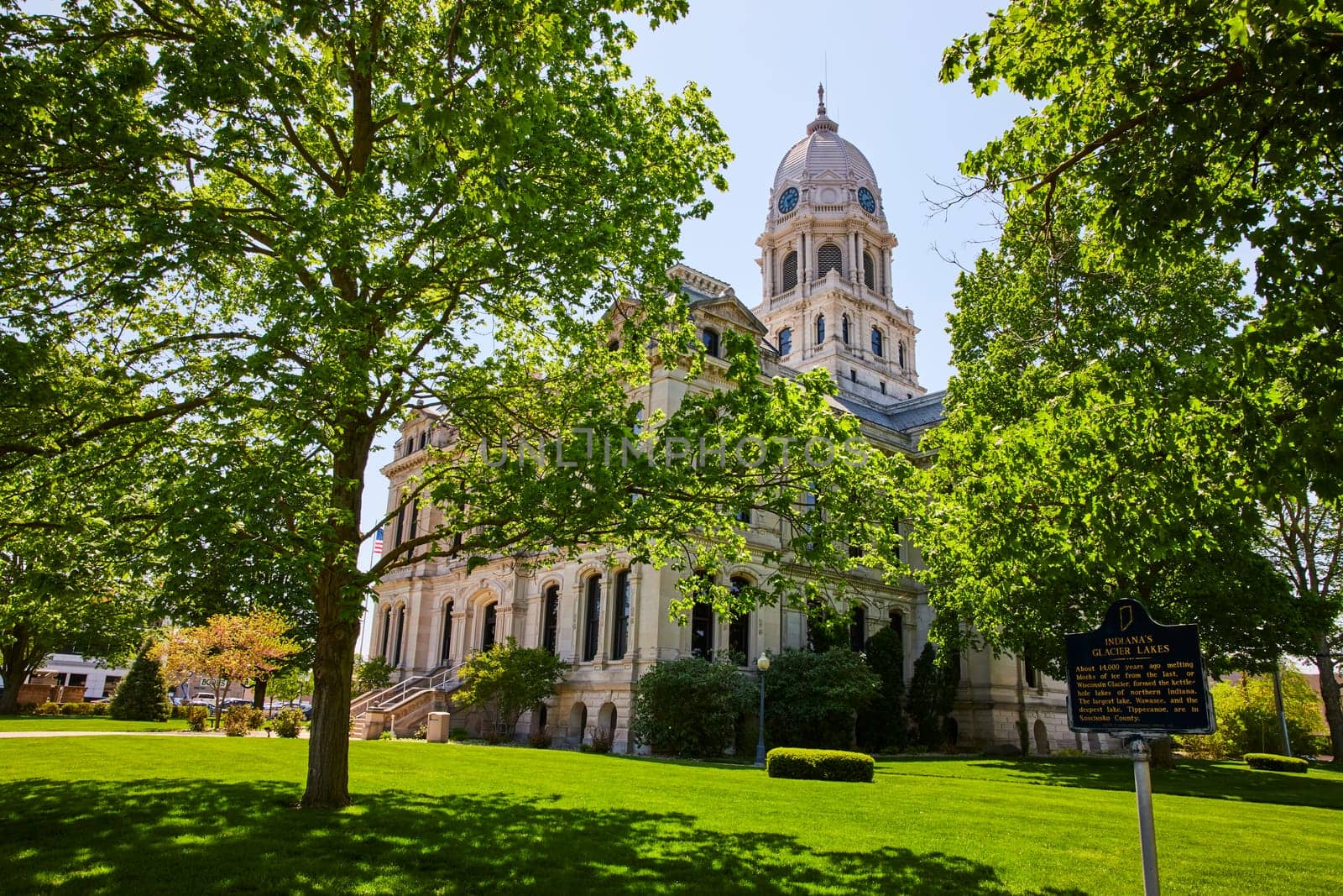 Historic Kosciusko County Courthouse in Warsaw, Indiana, surrounded by lush greenery under a clear blue sky.