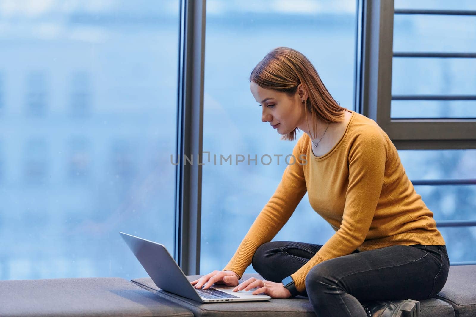 A businesswoman utilizes her laptop while seated by the window of a large corporate building, offering a picturesque view of the city skyline as her backdrop. by dotshock