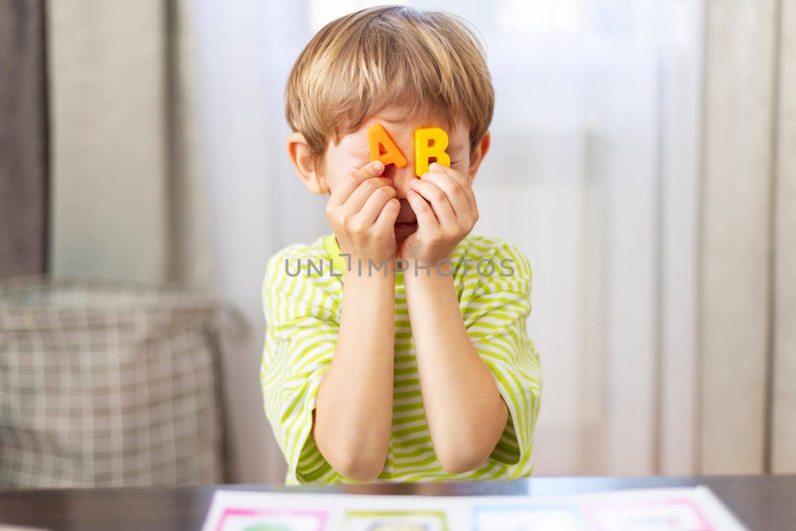 A young boy in a striped shirt engages in playful learning by covering his eyes with colorful alphabet letters, showcasing a moment of joy and early education in a home setting