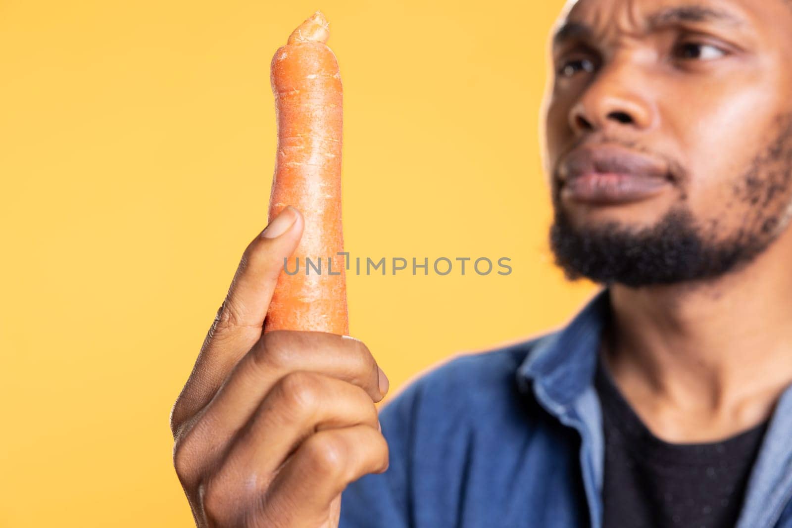African american guy in doubt inspecting an organic carrot, looking intensely at additives free freshly harvested vegetable. Young person examining natural locally grown veggie, zero waste.