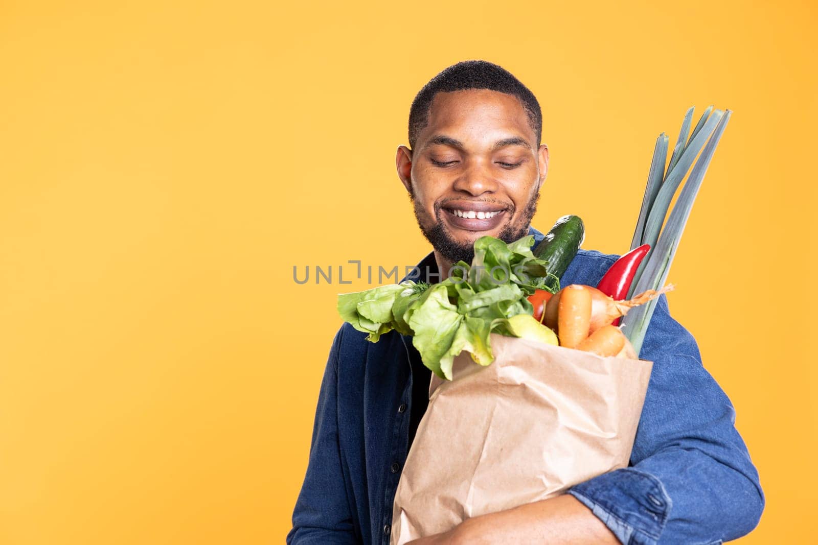 Positive enthusiastic guy smiling at his ethically sourced groceries, feeling pleased with his farmers market shopping session. Vegan person carrying his paper bag full of natural organic produce.
