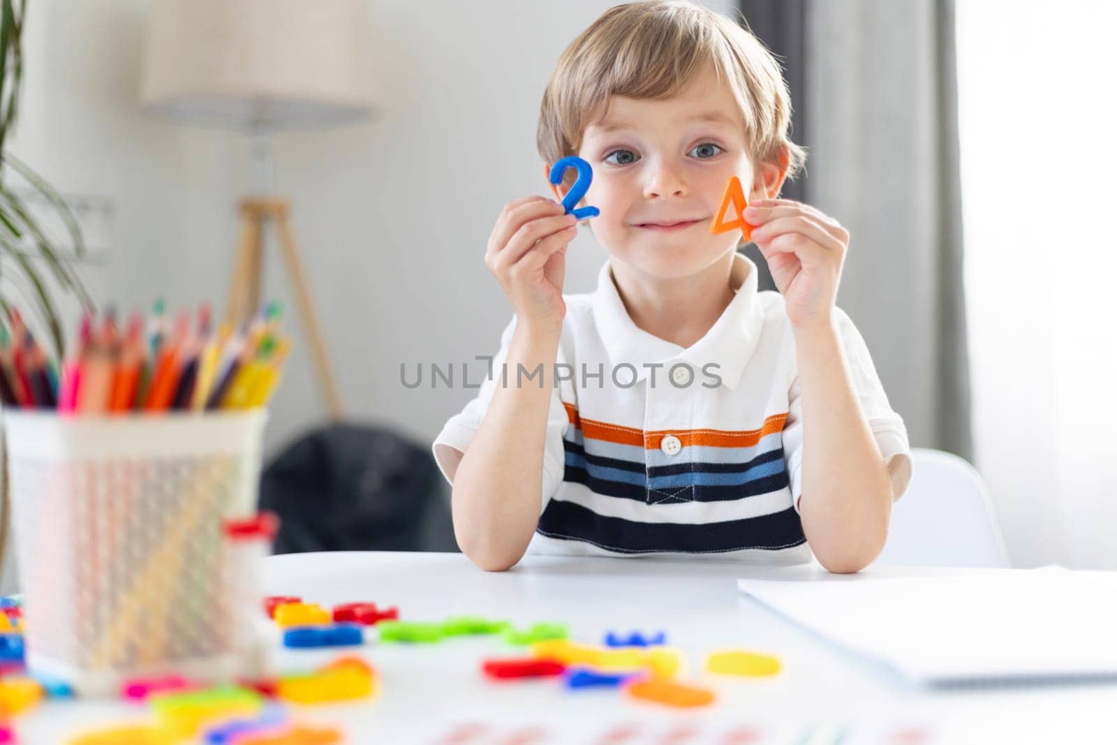 A young boy smiles at the camera while holding colorful geometric shapes, demonstrating a cheerful moment in a homeschooling environment.