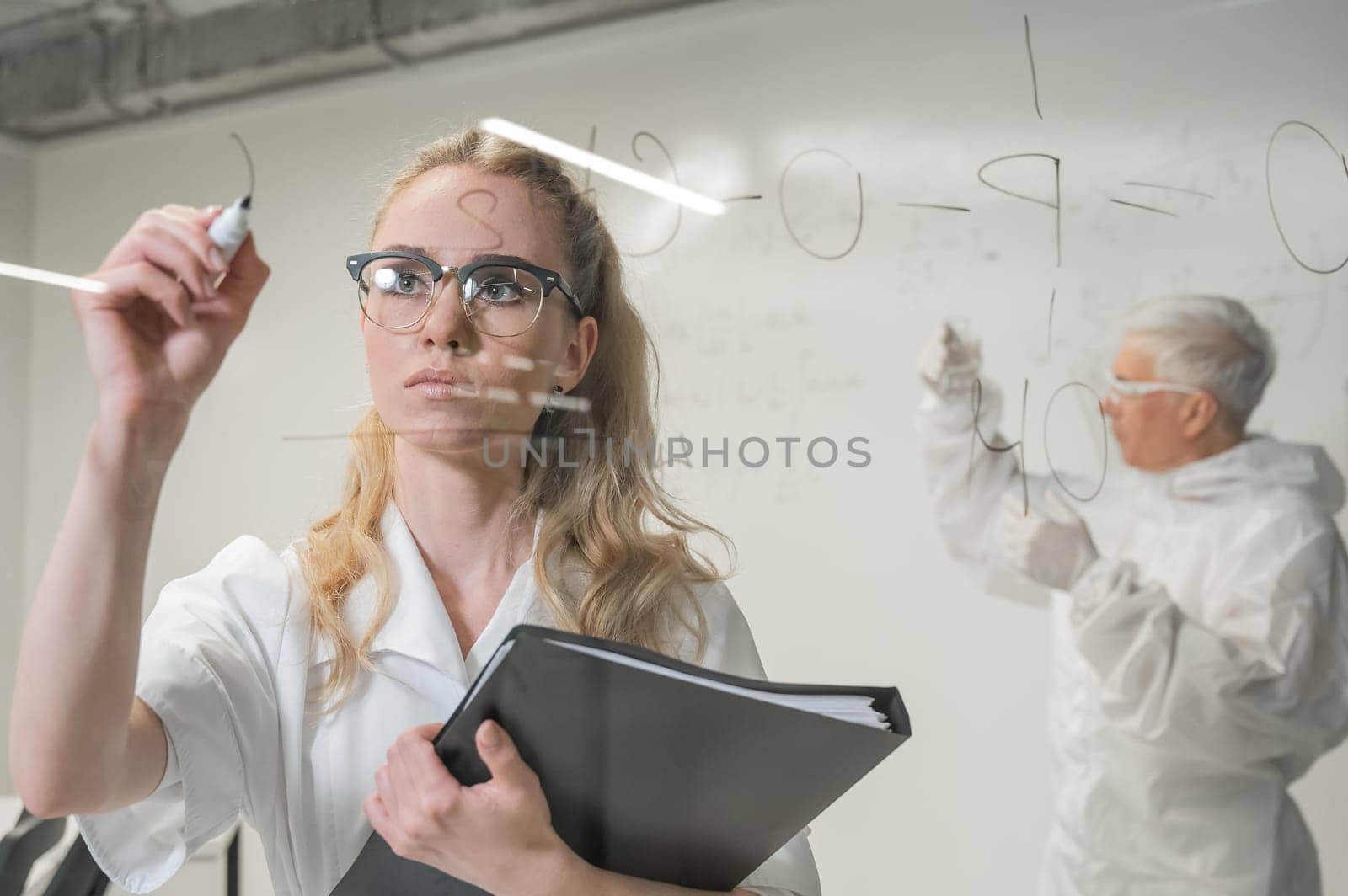 A woman chemist writes a formula on glass. An elderly Caucasian man in a protective suit is doing tests. by mrwed54