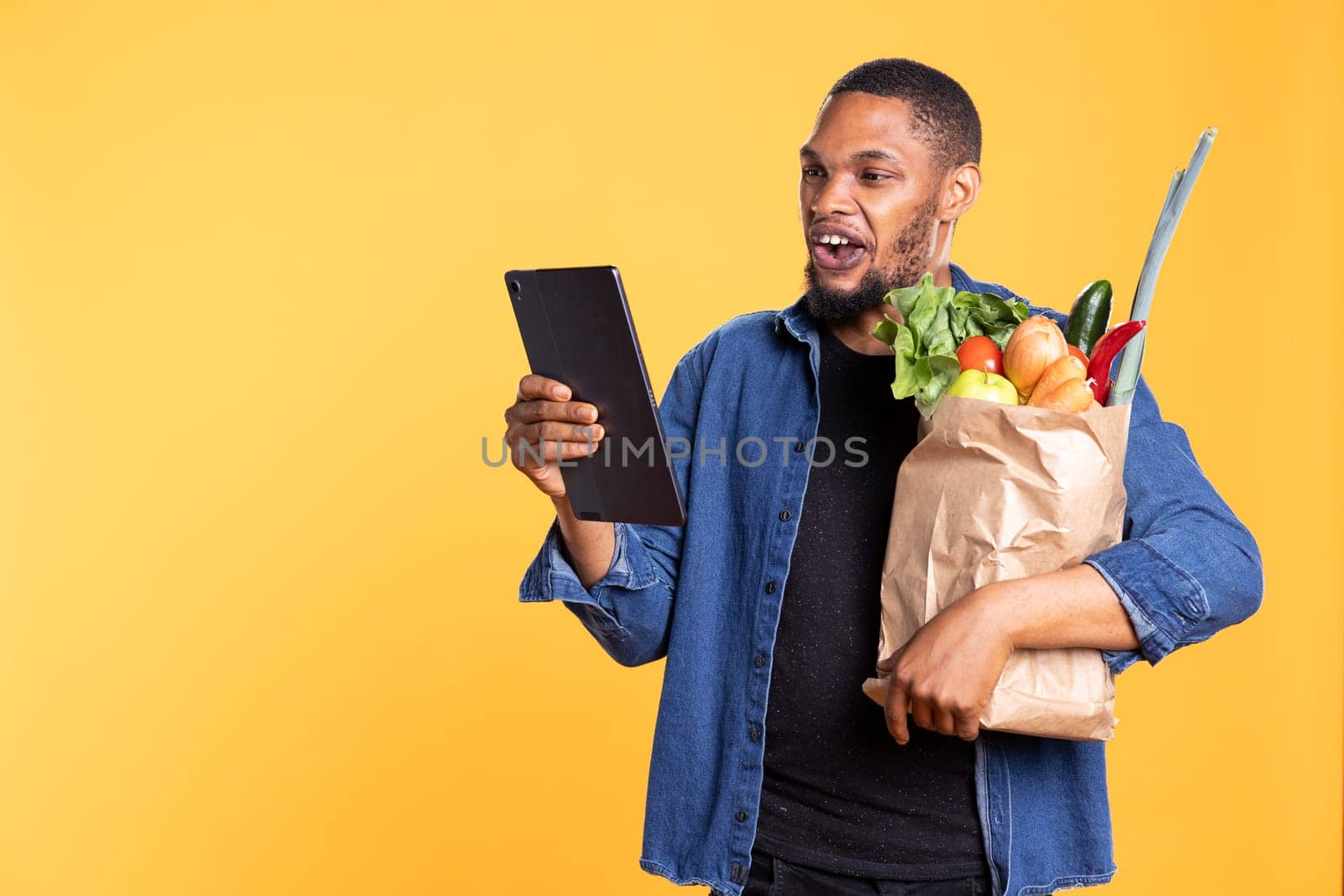 African american man checks his social media texts in studio, carrying a paper bag full of fruits and veggies from the local supermarket store. Confident relaxed guy having fun browsing the internet.