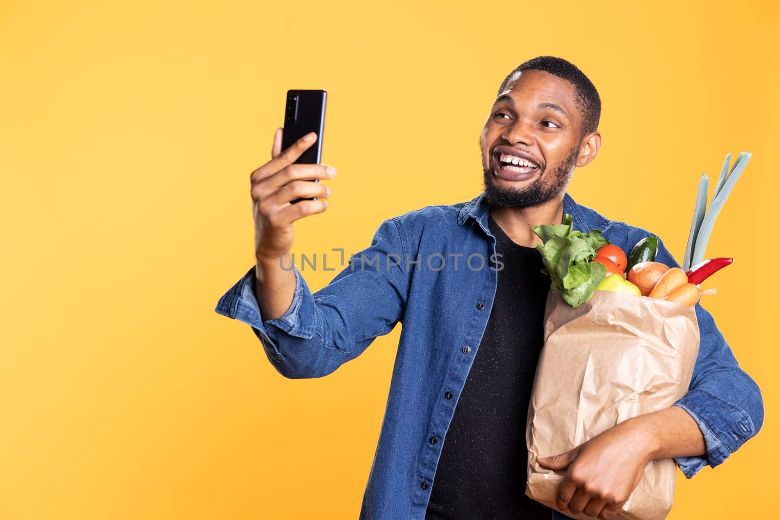 Smiling positive guy taking a picture with his bag of fresh produce, feels pleased with ethically sourced goods. Optimistic friendly person takes photos on his mobile phone app.