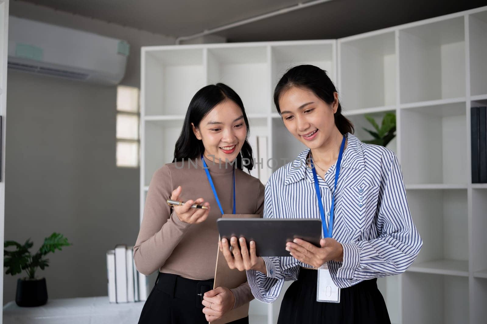 Two Asian businesswomen discussing work using a tablet in a modern office. Concept of teamwork and professional collaboration by wichayada