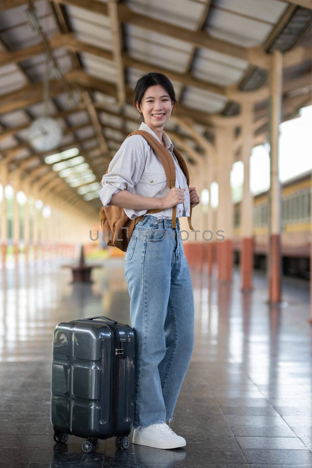 Happy young Asian female tourist carrying a backpack, holding a camera, preparing to wait for the train at the train station waiting for her vacation trip. by wichayada