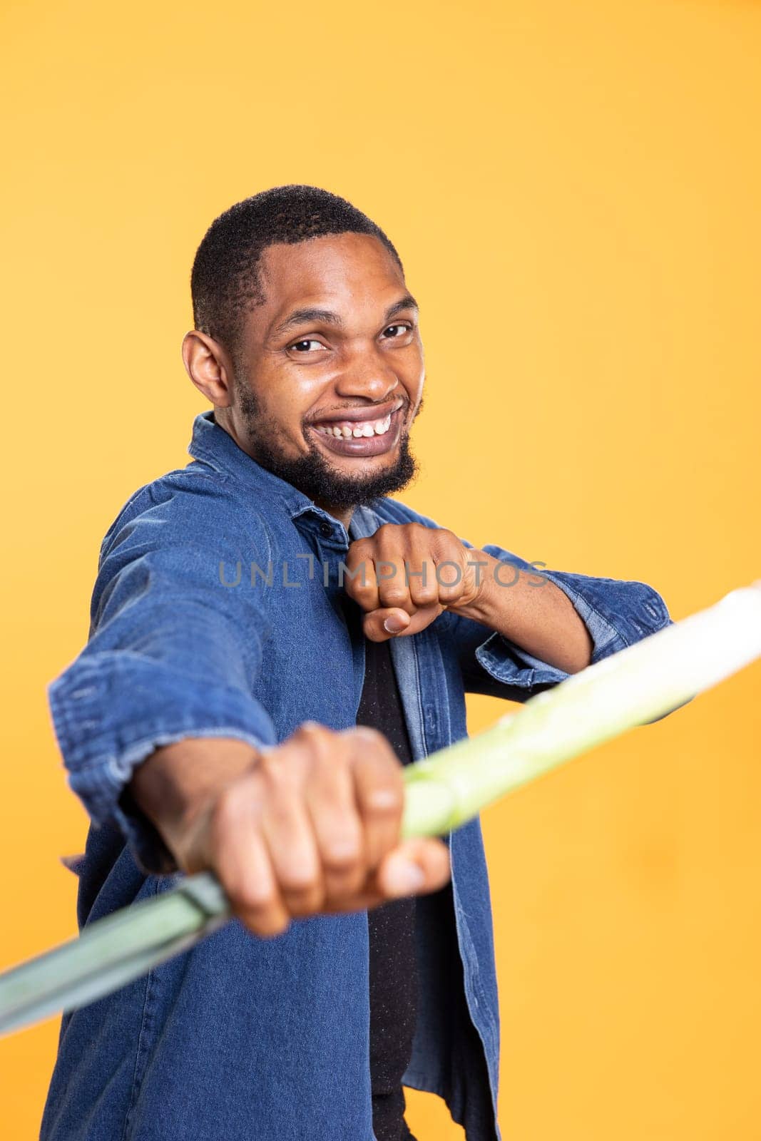 African american adult mimicking a combat move with a leek, holding produce as a sword against yellow background. Confident silly guy acting silly with locally grown green onion.