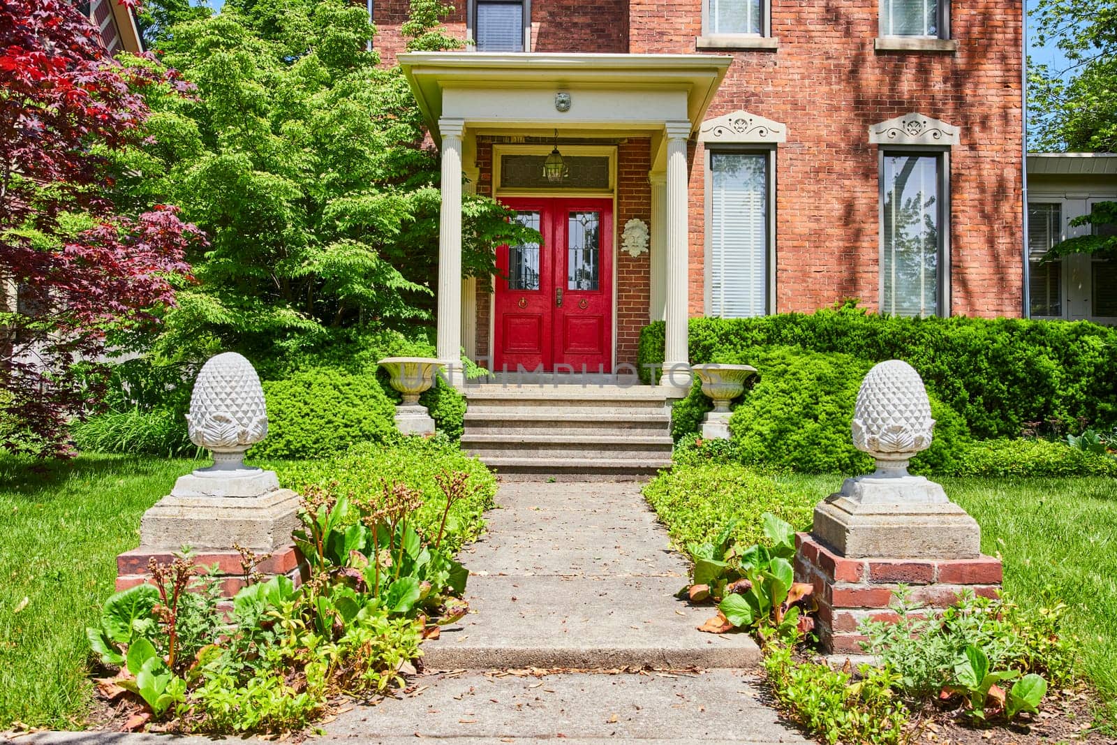 Sunny day at a classic brick home in Fort Wayne with vibrant red doors and lush greenery, embodying warmth and welcome.