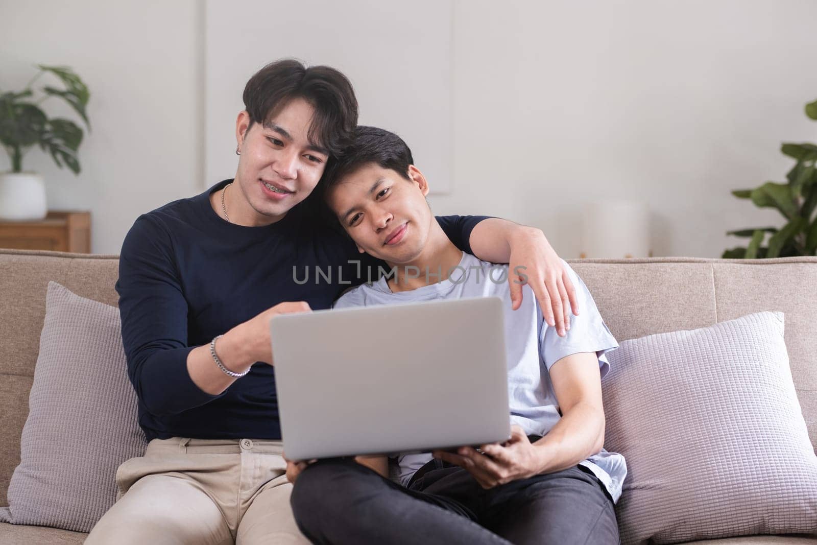 A gay couple spends their free time sitting on a laptop doing activities together on a holiday in the living room. by wichayada