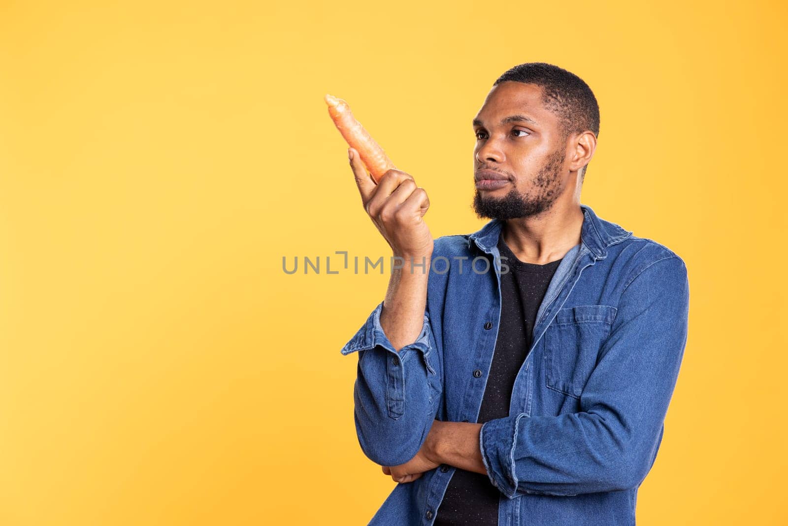 Happy charismatic person admiring ethically sourced carrots in studio, enjoying vegan nutrition and healthy eating. African american joyful guy smiling at organic ripe vegetables.