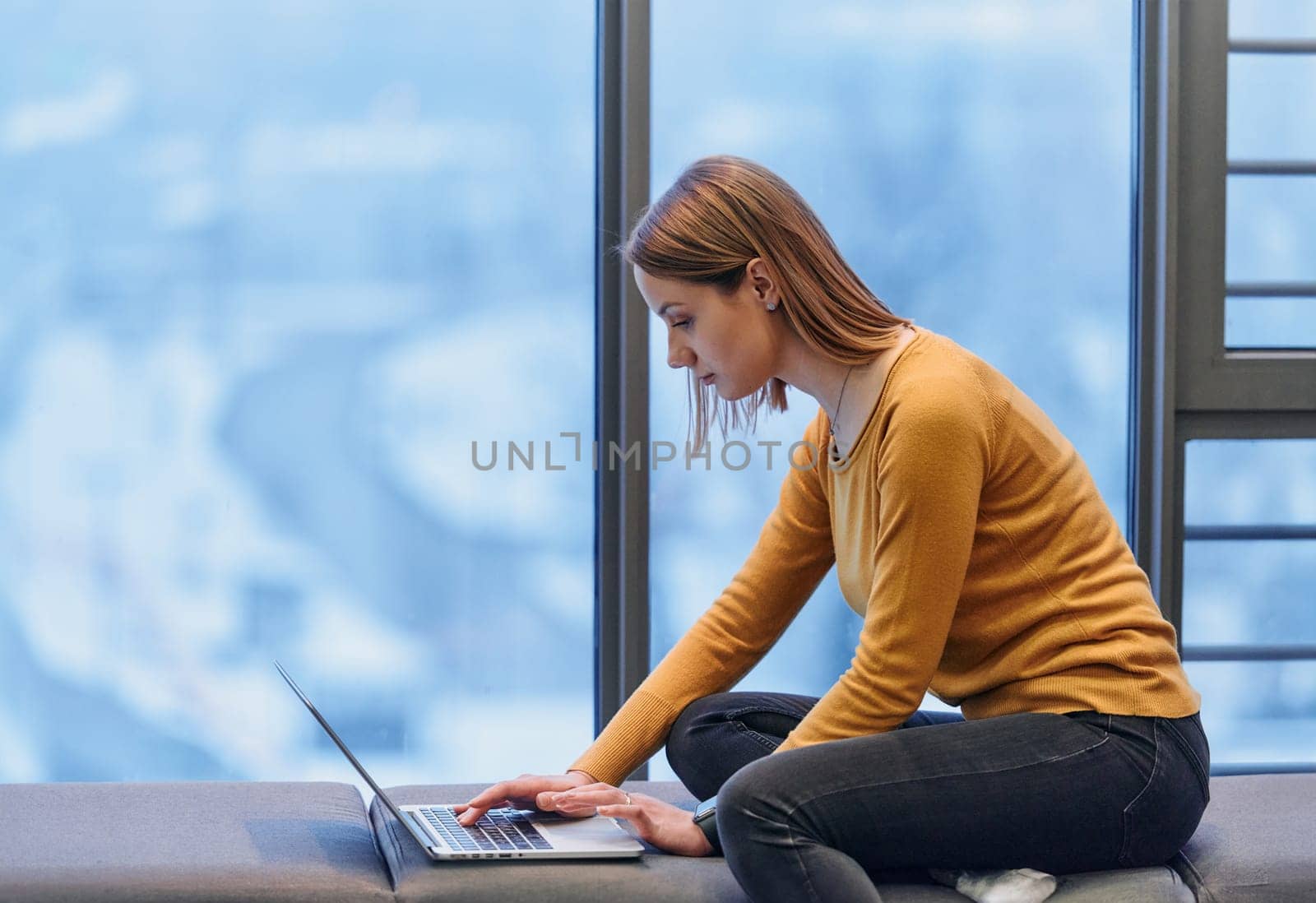 A businesswoman utilizes her laptop while seated by the window of a large corporate building, offering a picturesque view of the city skyline as her backdrop