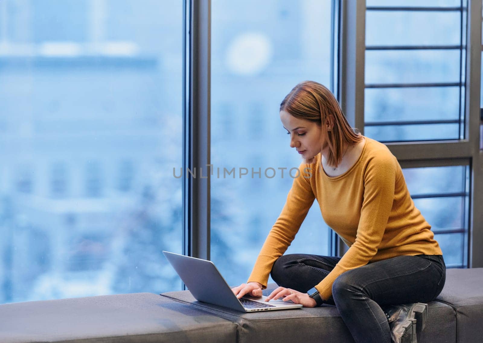 A businesswoman utilizes her laptop while seated by the window of a large corporate building, offering a picturesque view of the city skyline as her backdrop