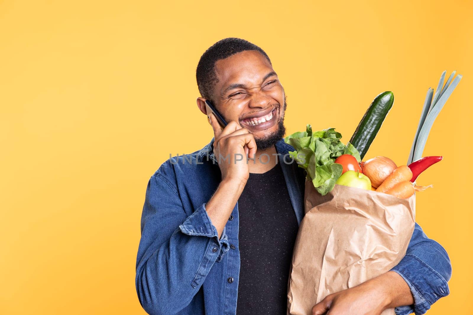 African american excited guy chatting on phone call and holds bag of groceries, discussing with his friends on remote connection line. and Carrying ethically sourced produce in studio.