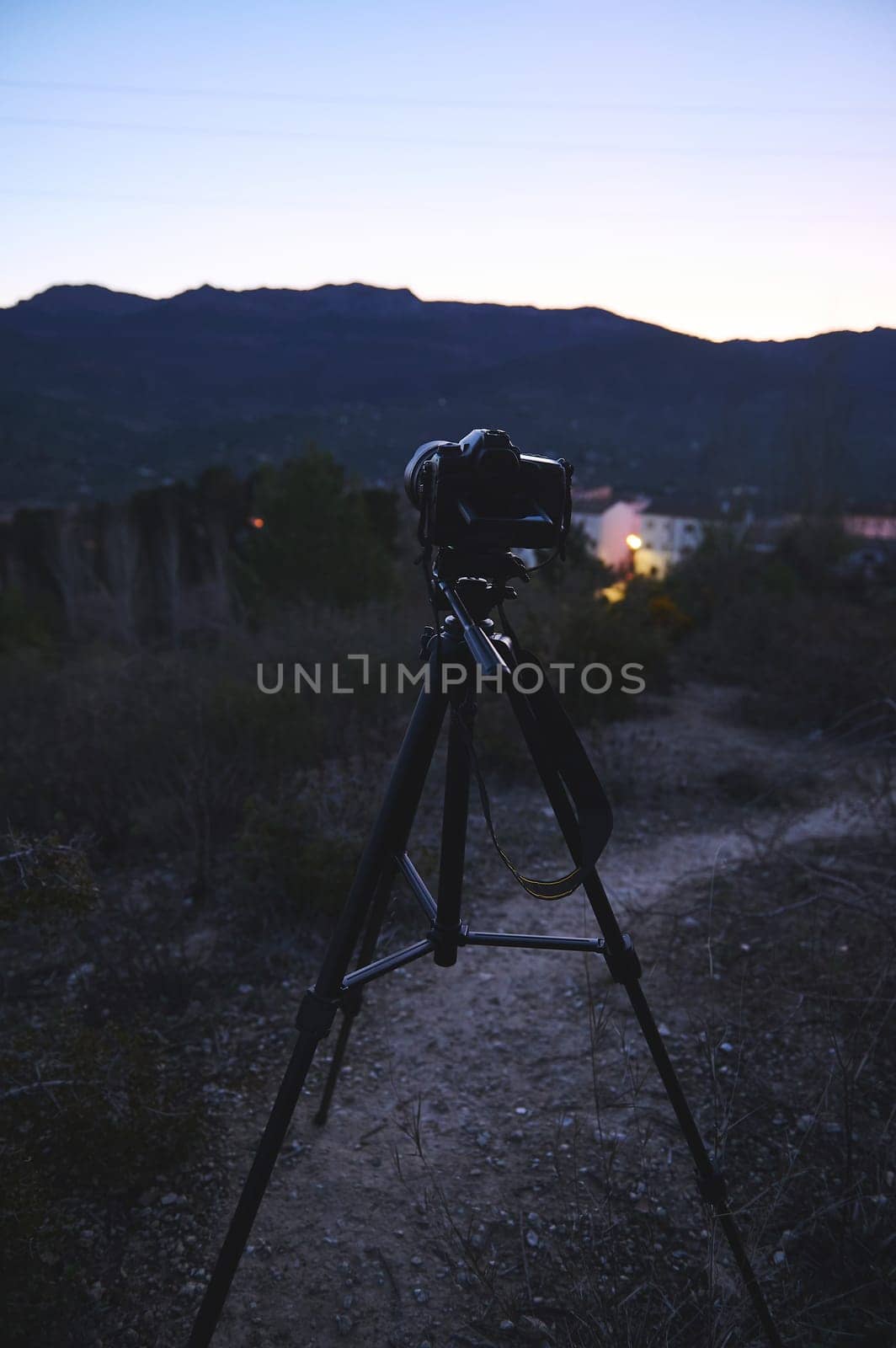 World photography day concept. Vertical shot of modern digital camera placed on tripod in nature outdoors, ready for capturing steady pictures or video of a village in mountains at dawn