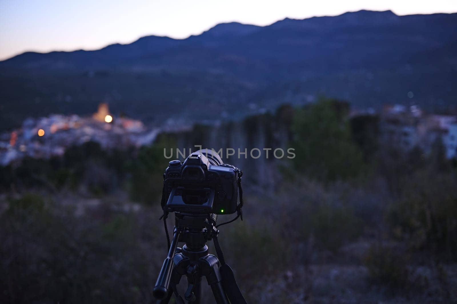 World photography day concept. Horizontal shot of modern digital camera placed on tripod in nature outdoors, ready for capturing steady pictures or video of a village in mountains at dawn