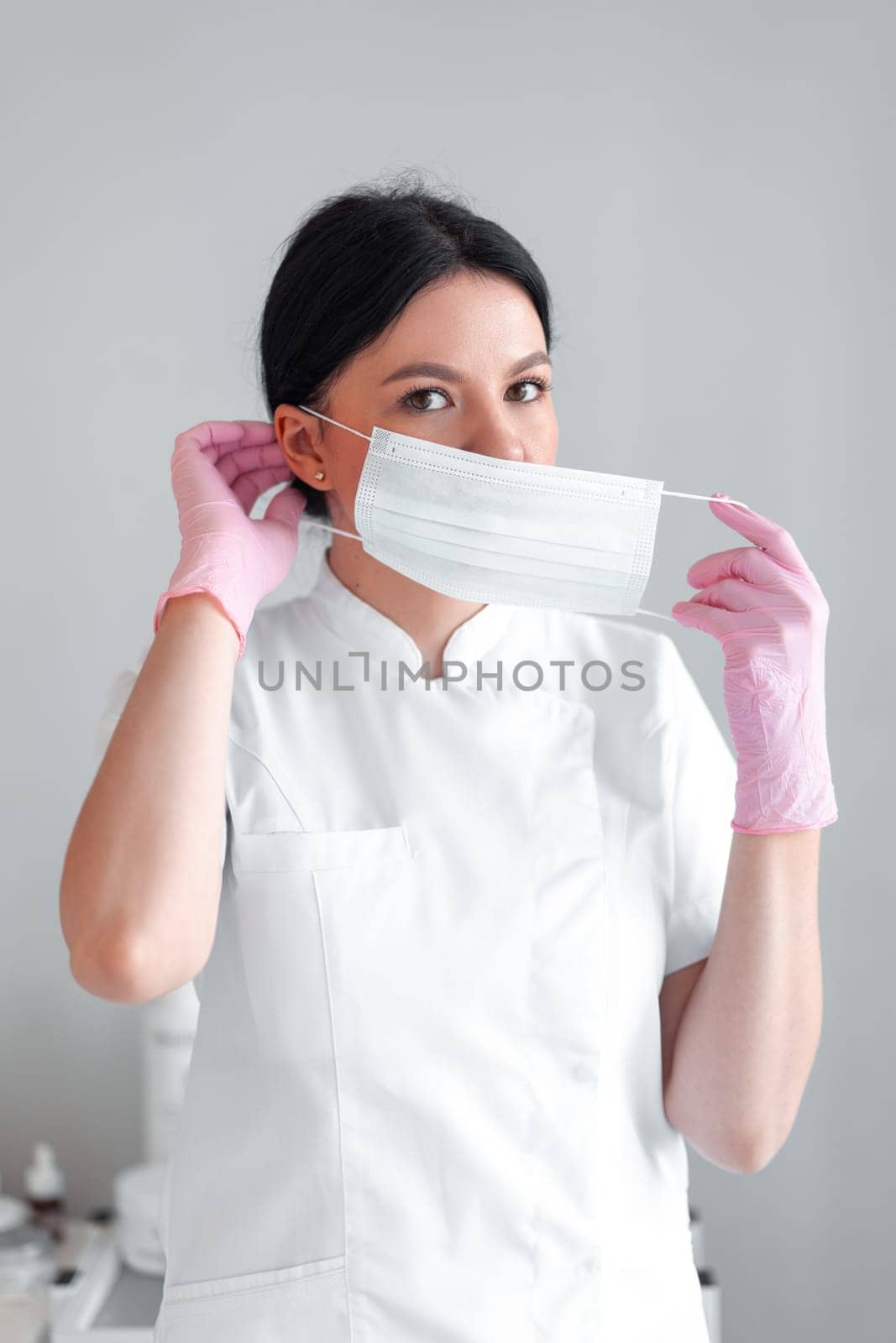 Female cosmetologist putting on mask before beauty treatment in clinic. Portrait of confident woman dermatologist in pink gloves and uniform working in hospital.