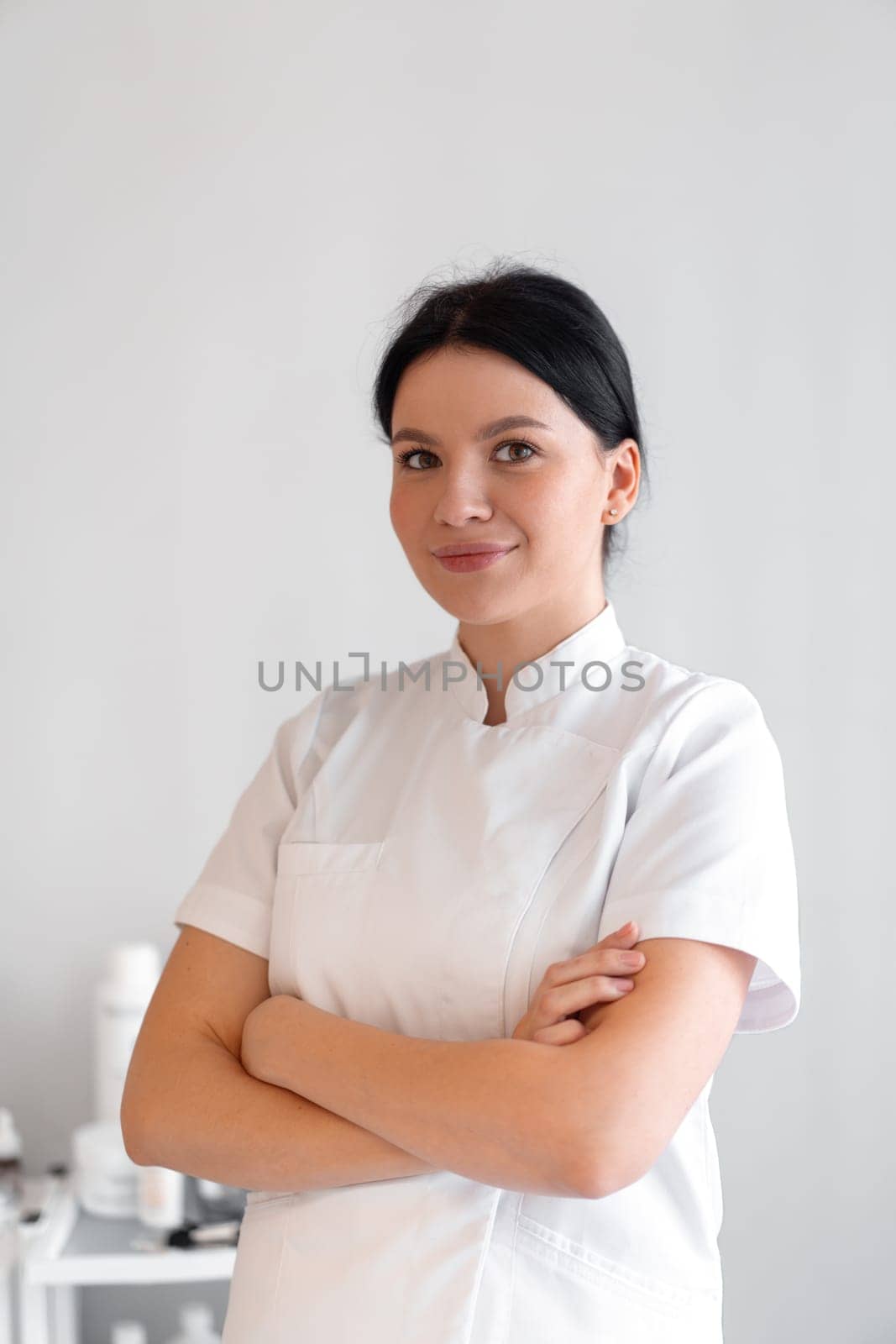 Smiling female cosmetologist in uniform standing arms crossed. Portrait of young woman doctor in white coat looking at camera at beauty salon.