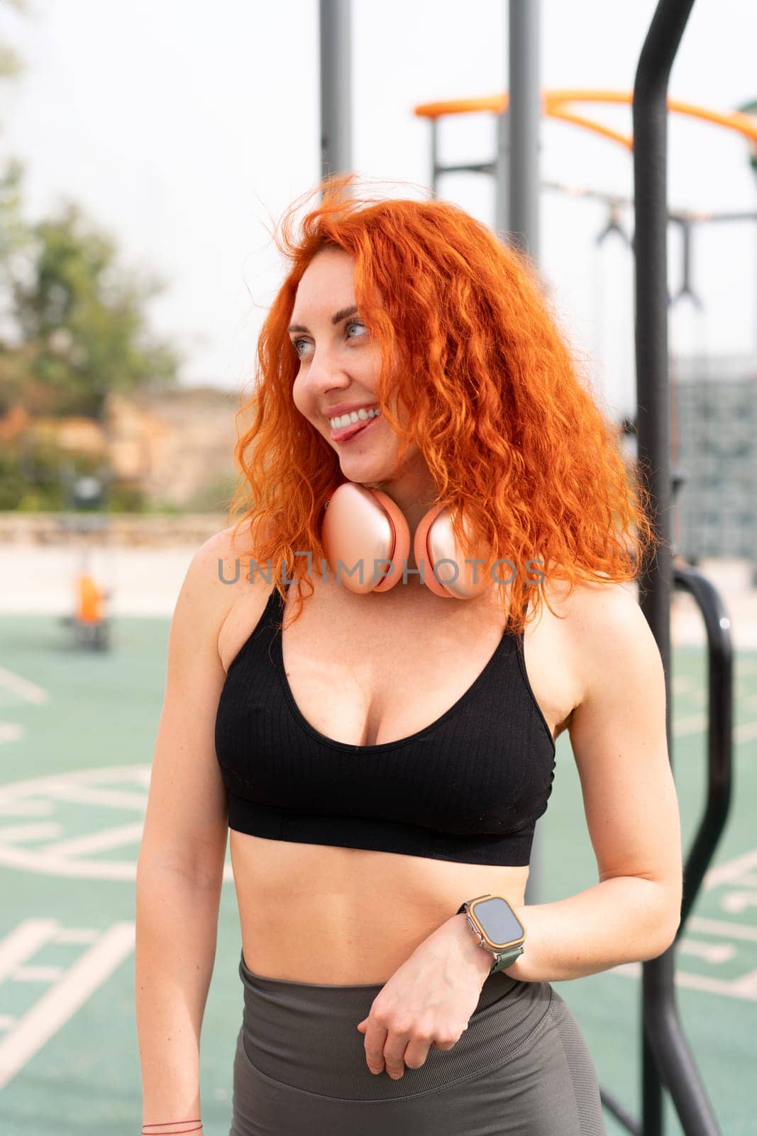 Funny sportswoman biting tongue and looking away. Smiling fit woman athlete with red hair in sportswear standing on outdoor gym park on sunny day.