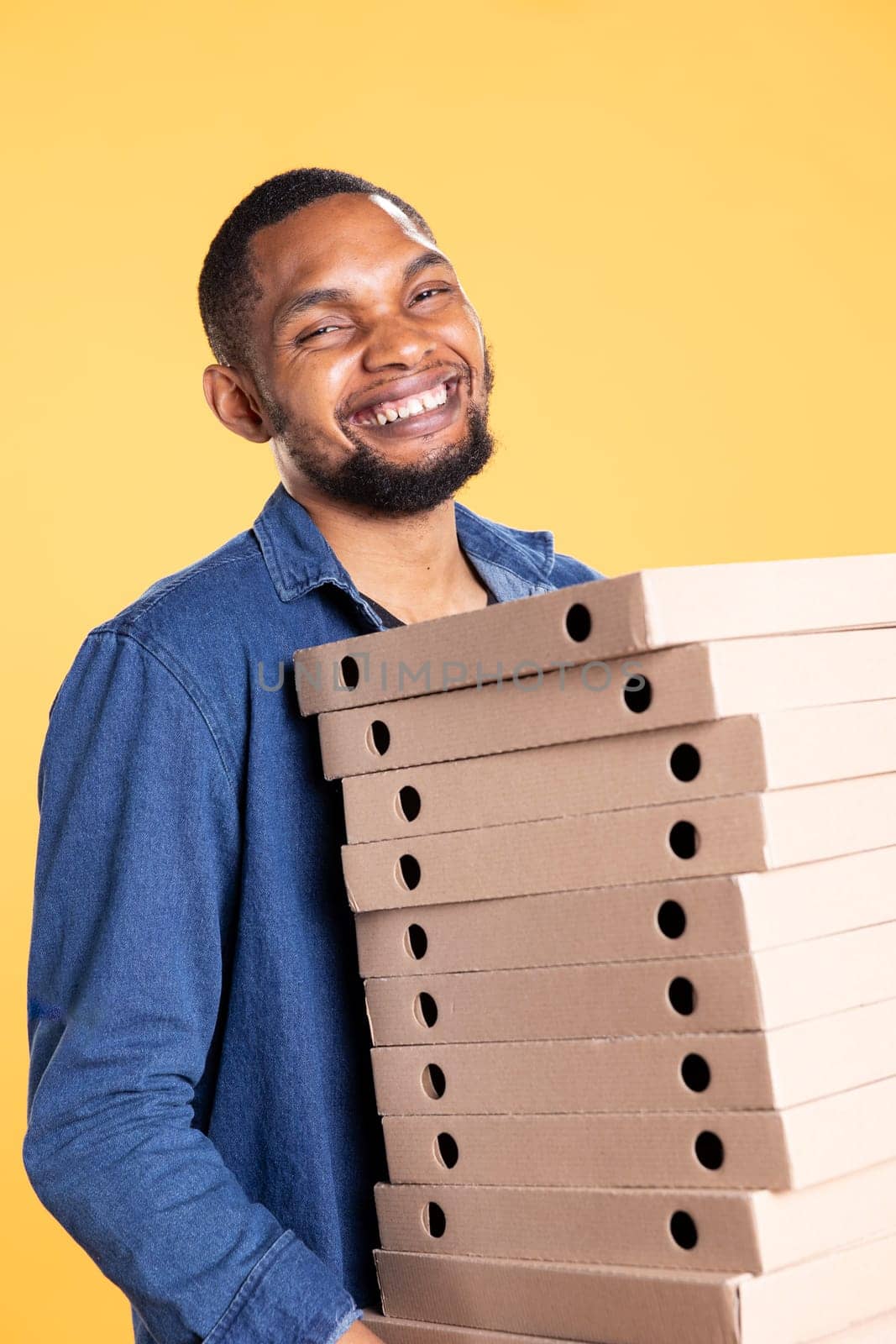 African american pizzeria deliveryman holding pile of pizza boxes to delivering food order to customer in studio. Friendly takeaway service worker carries takeout meal stack package.